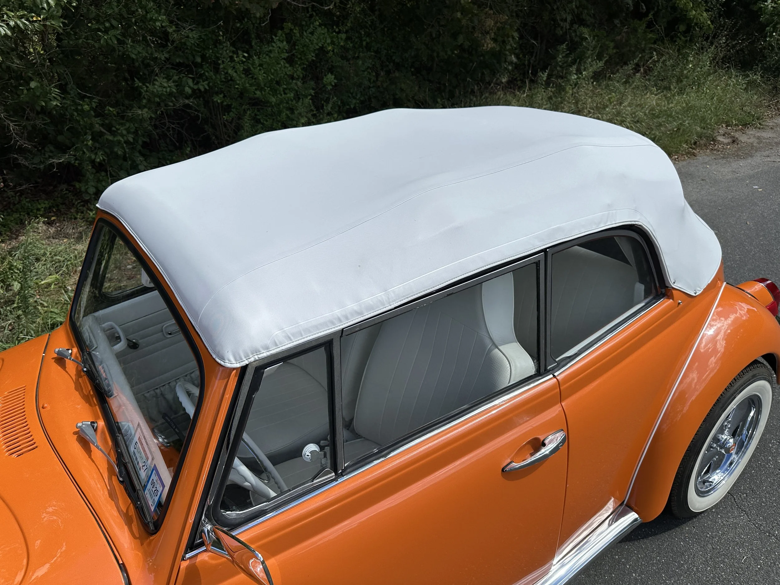 An orange vintage convertible car with a white soft top, parked on a road next to green bushes.
