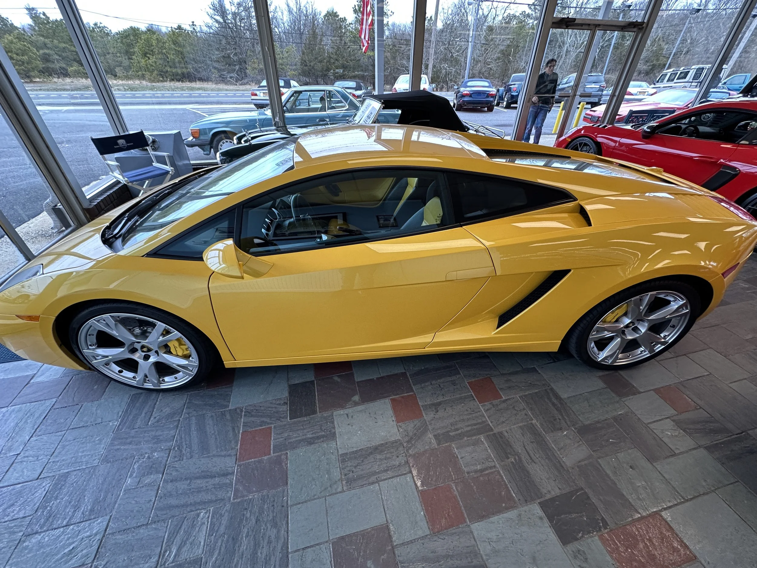 A yellow Lamborghini sports car displayed inside a showroom with large windows, surrounded by other cars and people outside.