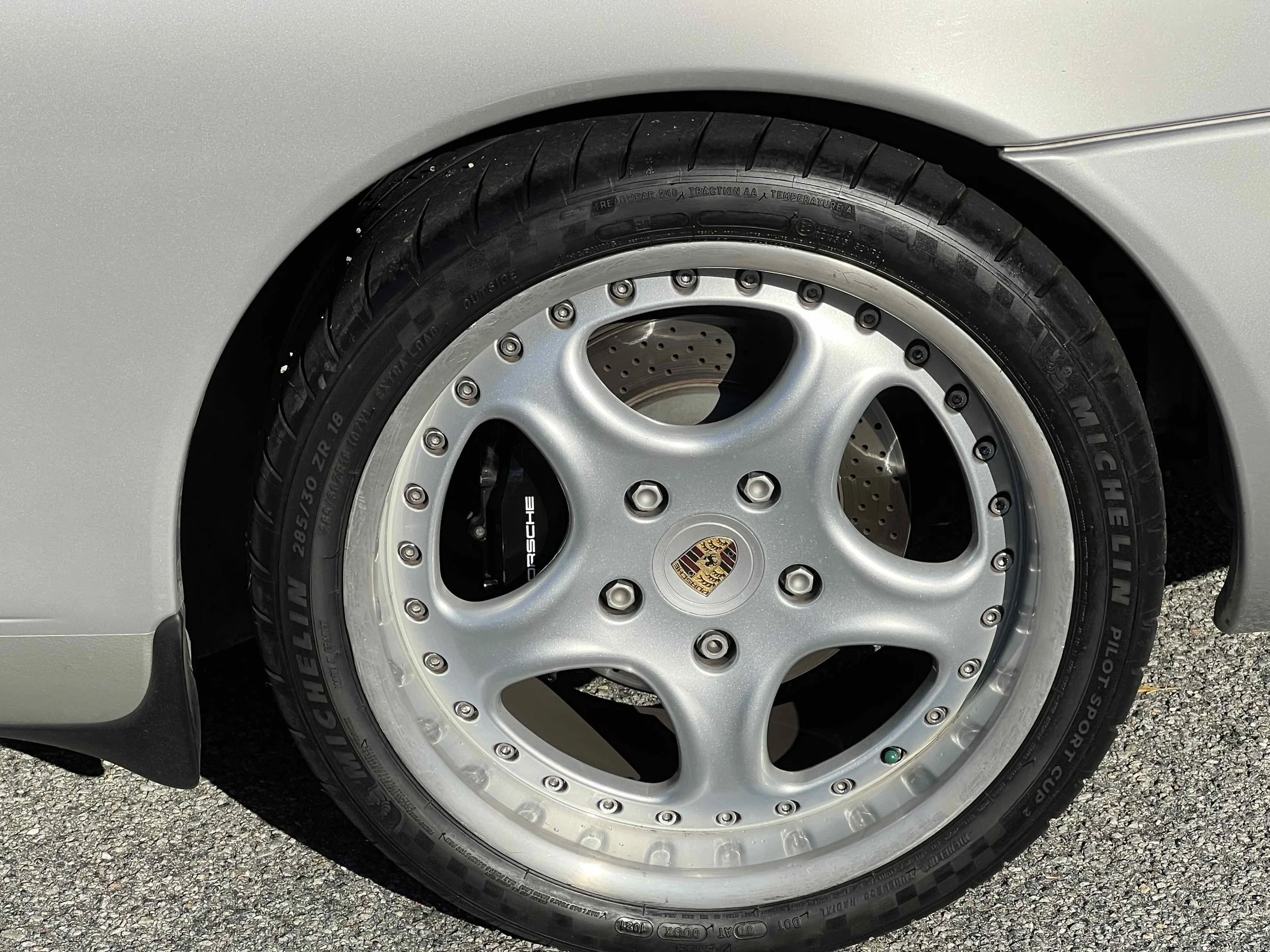 Close-up of a silver Porsche car wheel with a Michelin Pilot Sport Cup 2 tire, showing a black Porsche logo center cap with the Porsche crest, drilled brake rotor behind the wheel, and a black brake caliper.