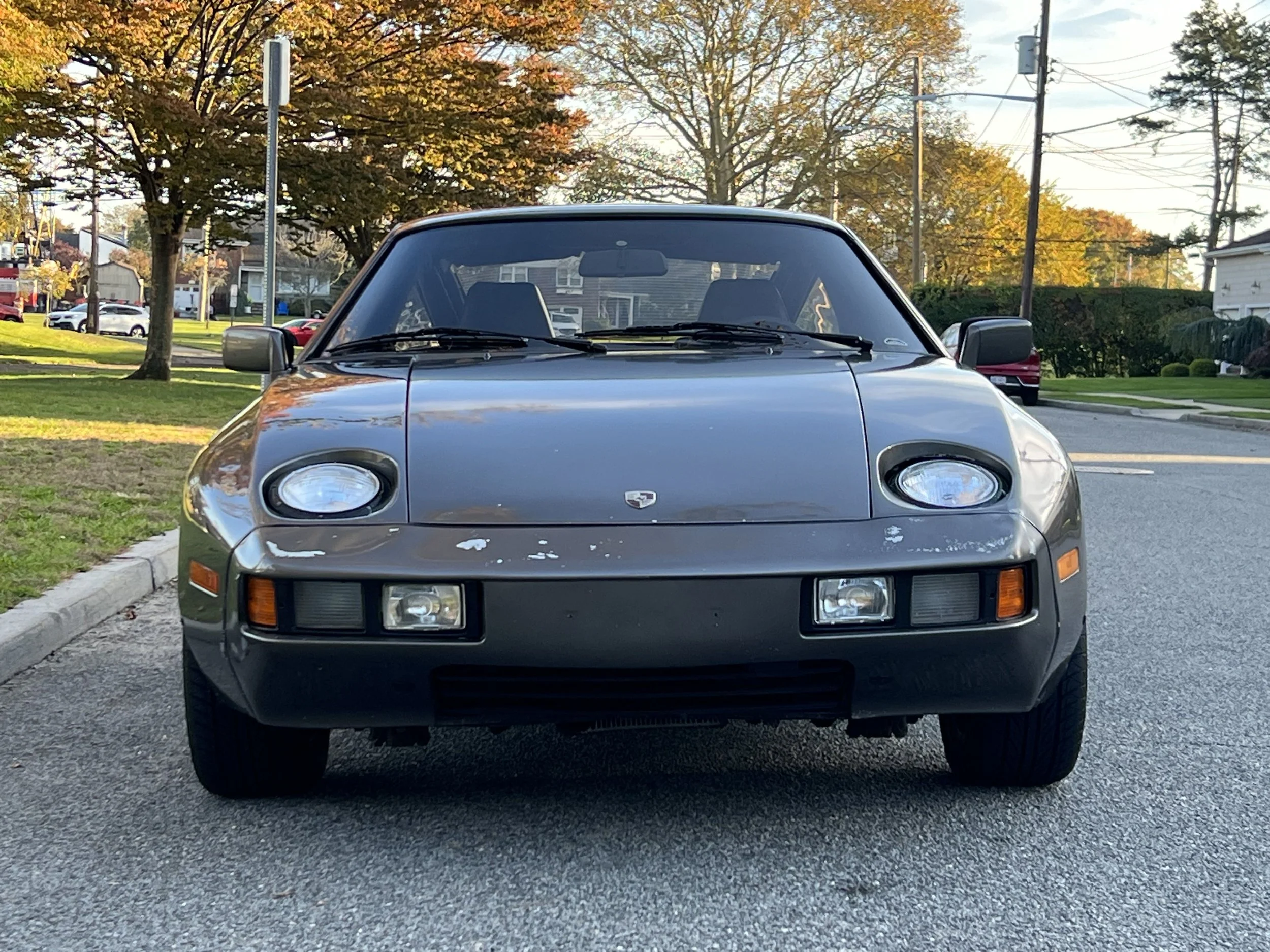 Front view of a gray vintage sports car with pop-up headlights parked on a suburban street during autumn.