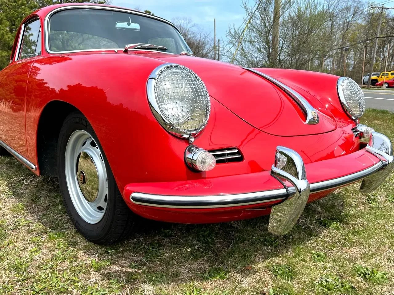 Red vintage Porsche 911 sports car parked on grass with trees, road, and cars in the background, featuring round headlights with protective grilles and chrome bumper guards.