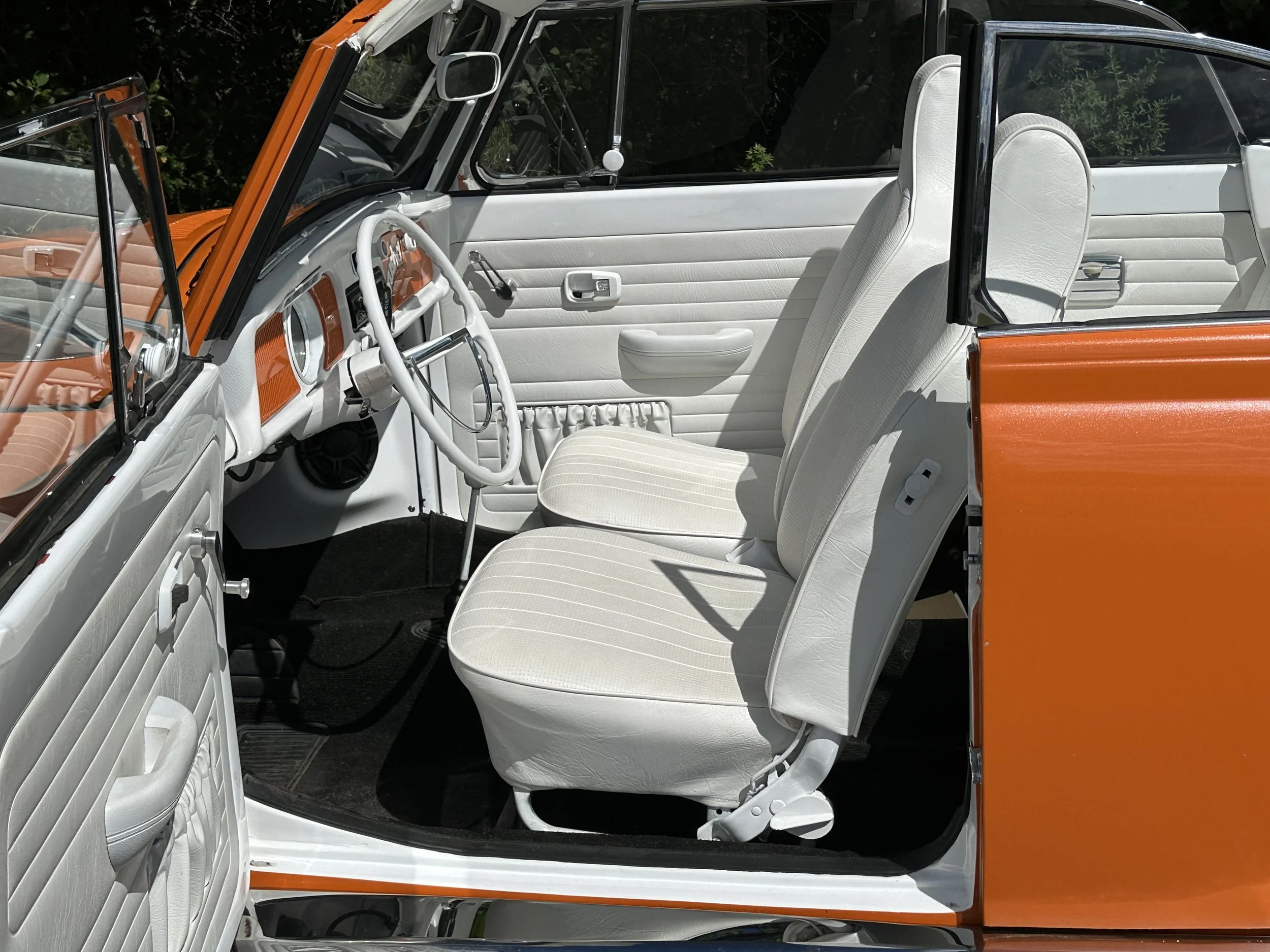 Interior of a vintage car with white seats and dashboard, orange exterior, and black carpet.