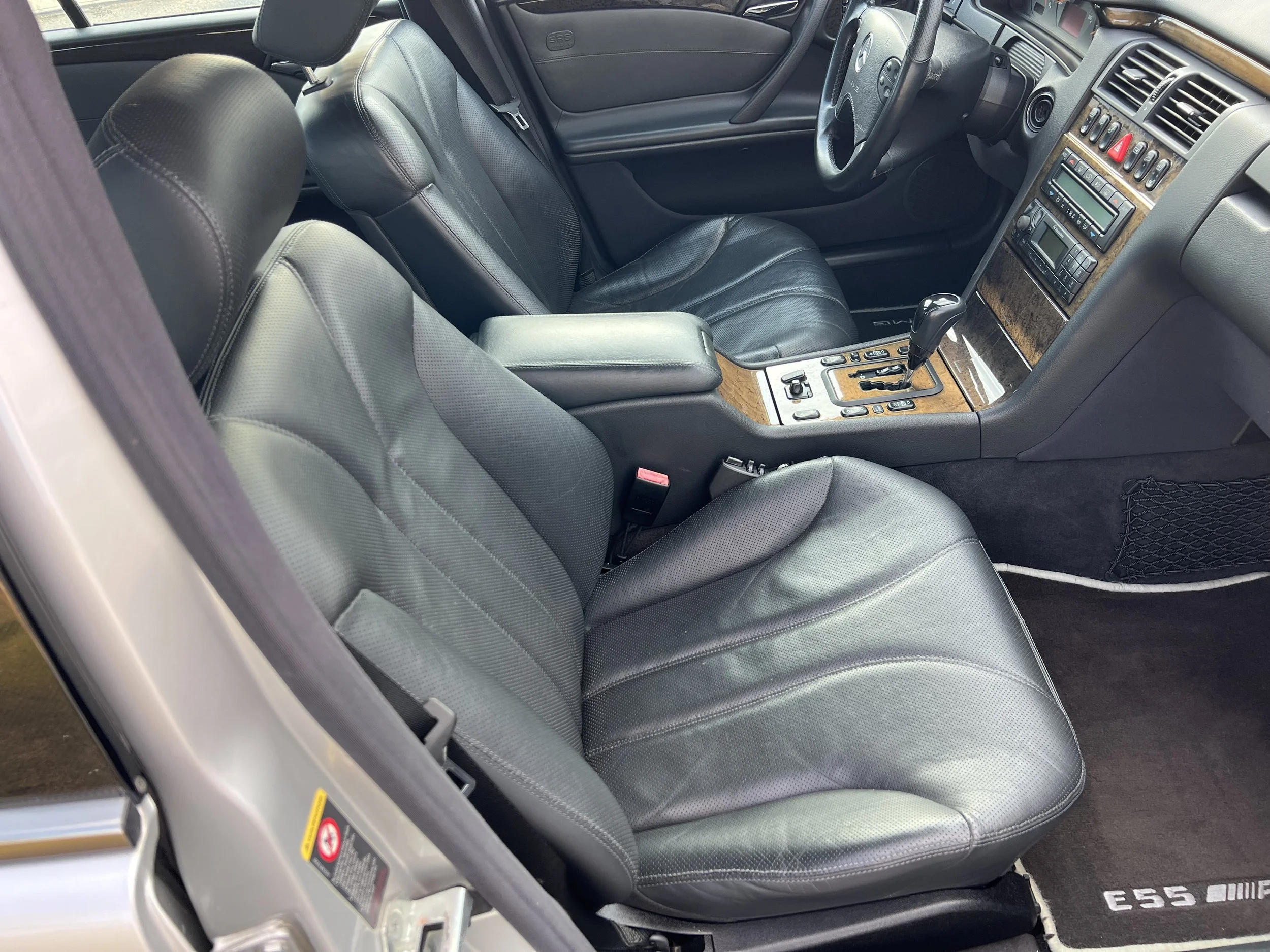 Interior view of a car showing the driver and passenger seats made of black leather, with wood trim detailing on the center console and dashboard, including controls, gear shift, and air vents.