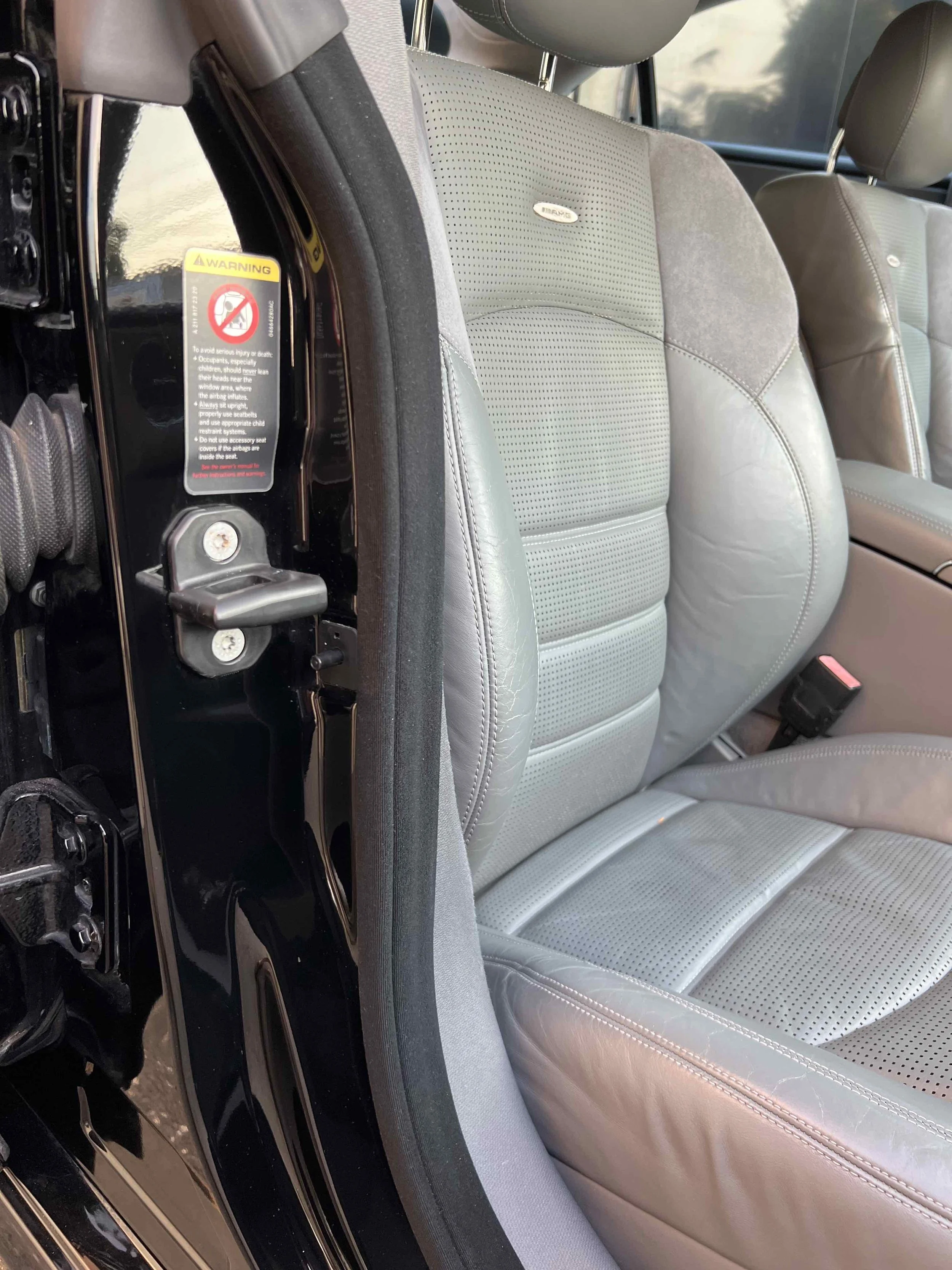 The image shows the front passenger seat interior of a vehicle, featuring light gray leather upholstery with perforated inserts, a seatbelt buckle, and part of the door frame with a warning label.
