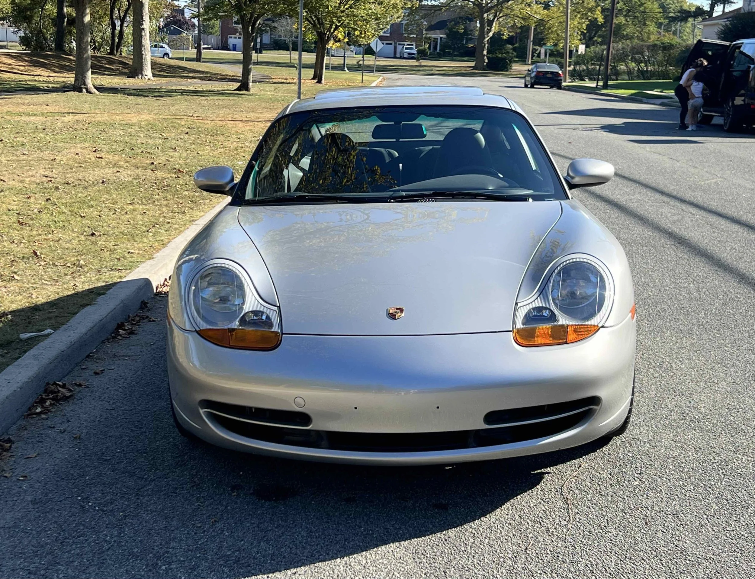 Front view of a silver Porsche sports car parked in a parking lot on a sunny day, with trees and people in the background.