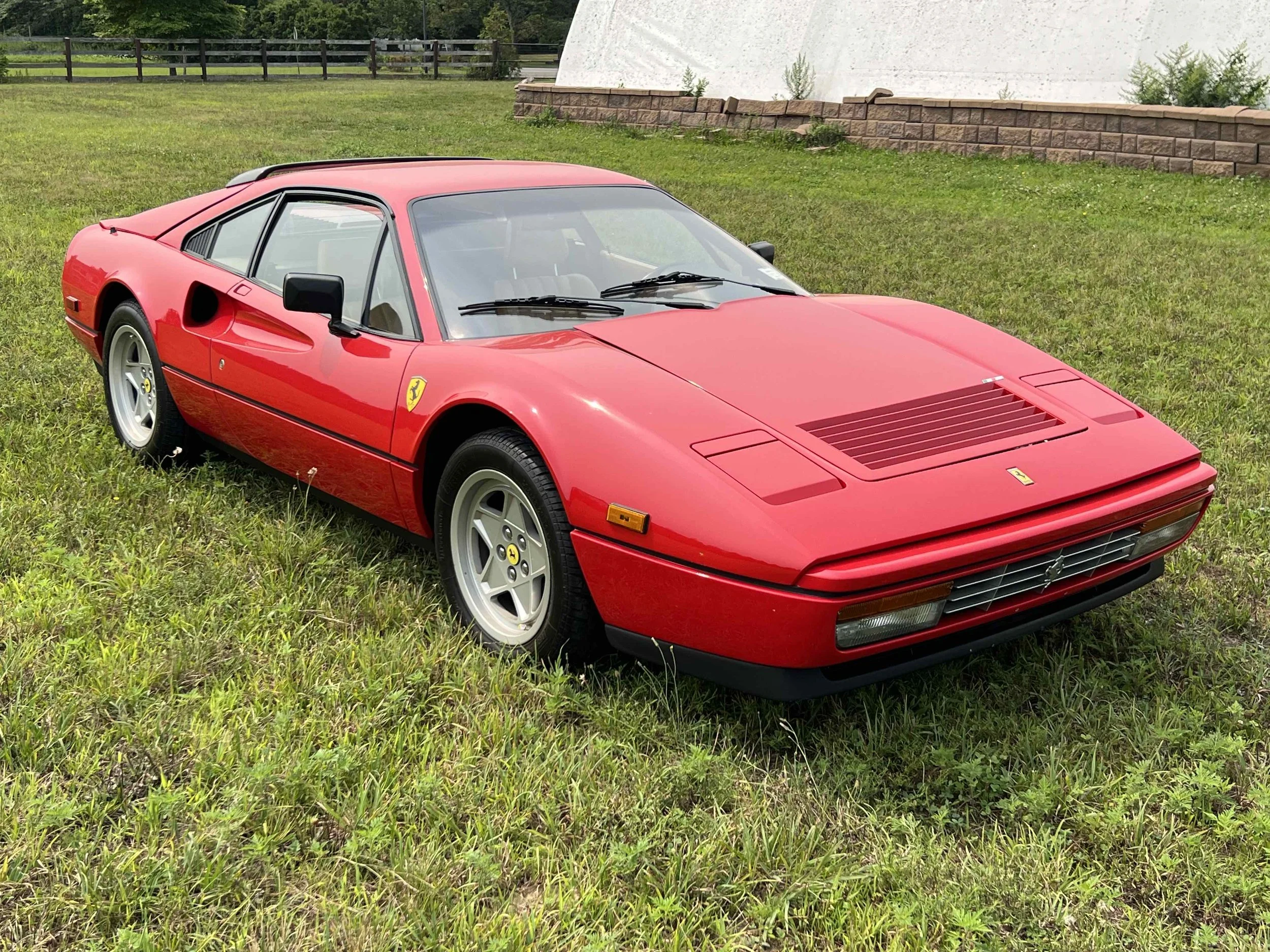 A red Ferrari sports car parked on green grass in front of a stone wall and white building.