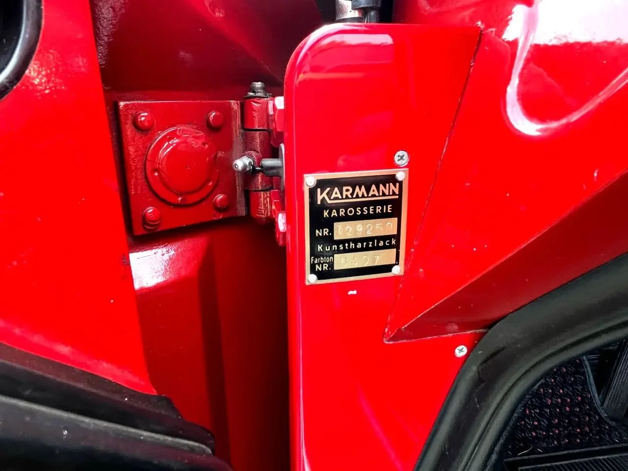Close-up of a red Karmann Ghia car's door hinge and identification plate with black background and gold lettering.