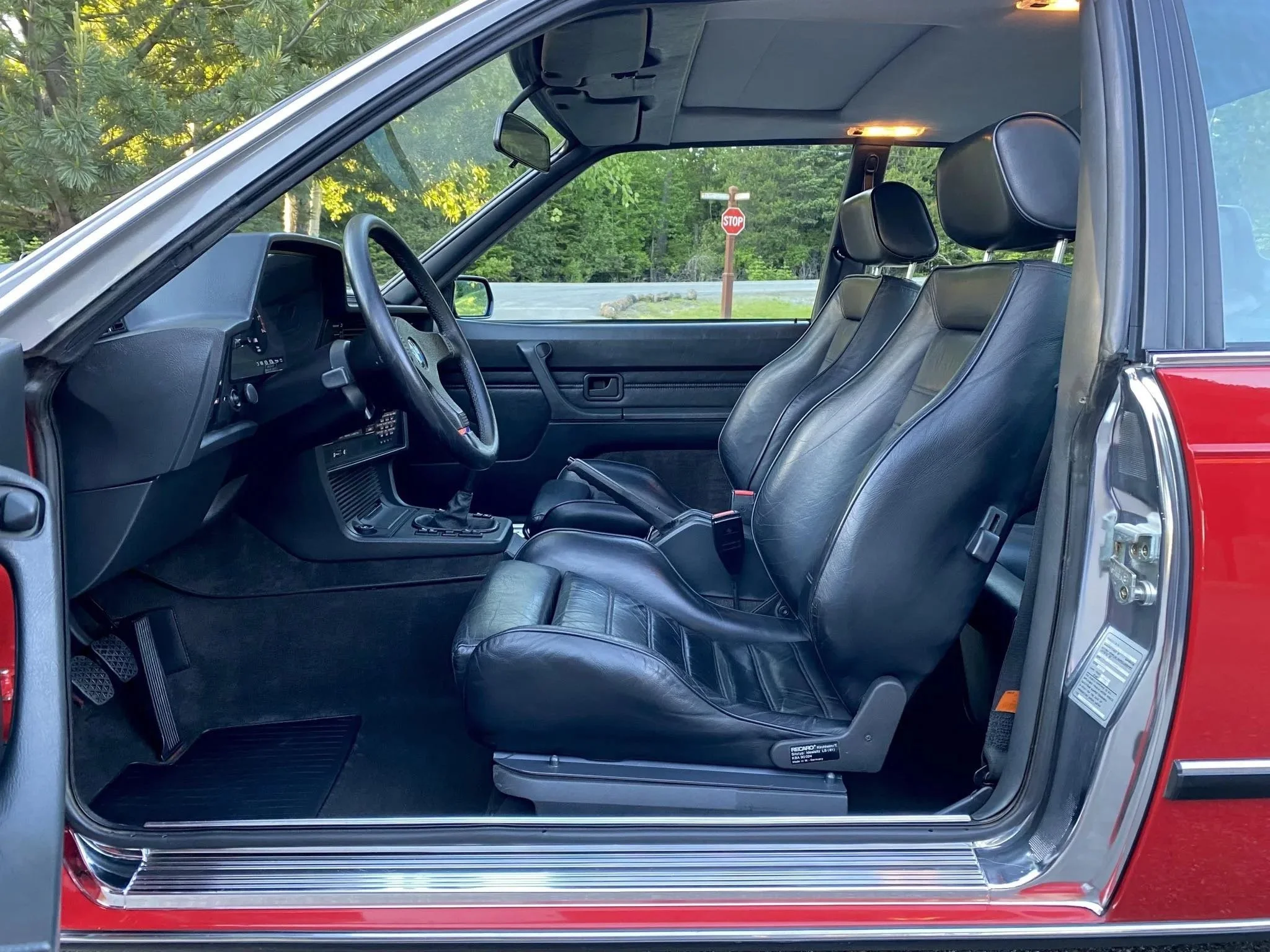 Interior of a vintage red car showing black leather front seats, dashboard, steering wheel, and inside door panel, with green foliage and a stop sign visible through the window.