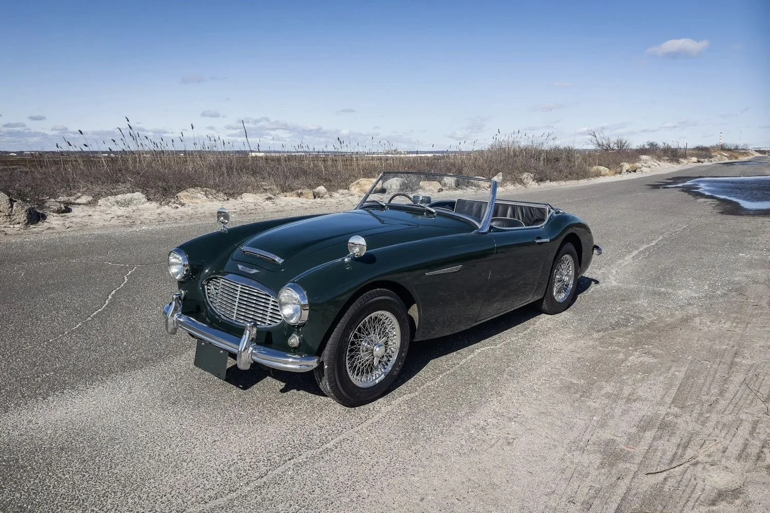 A vintage dark green convertible sports car parked on a sandy beach with a rocky shoreline and blue sky with some clouds in the background.