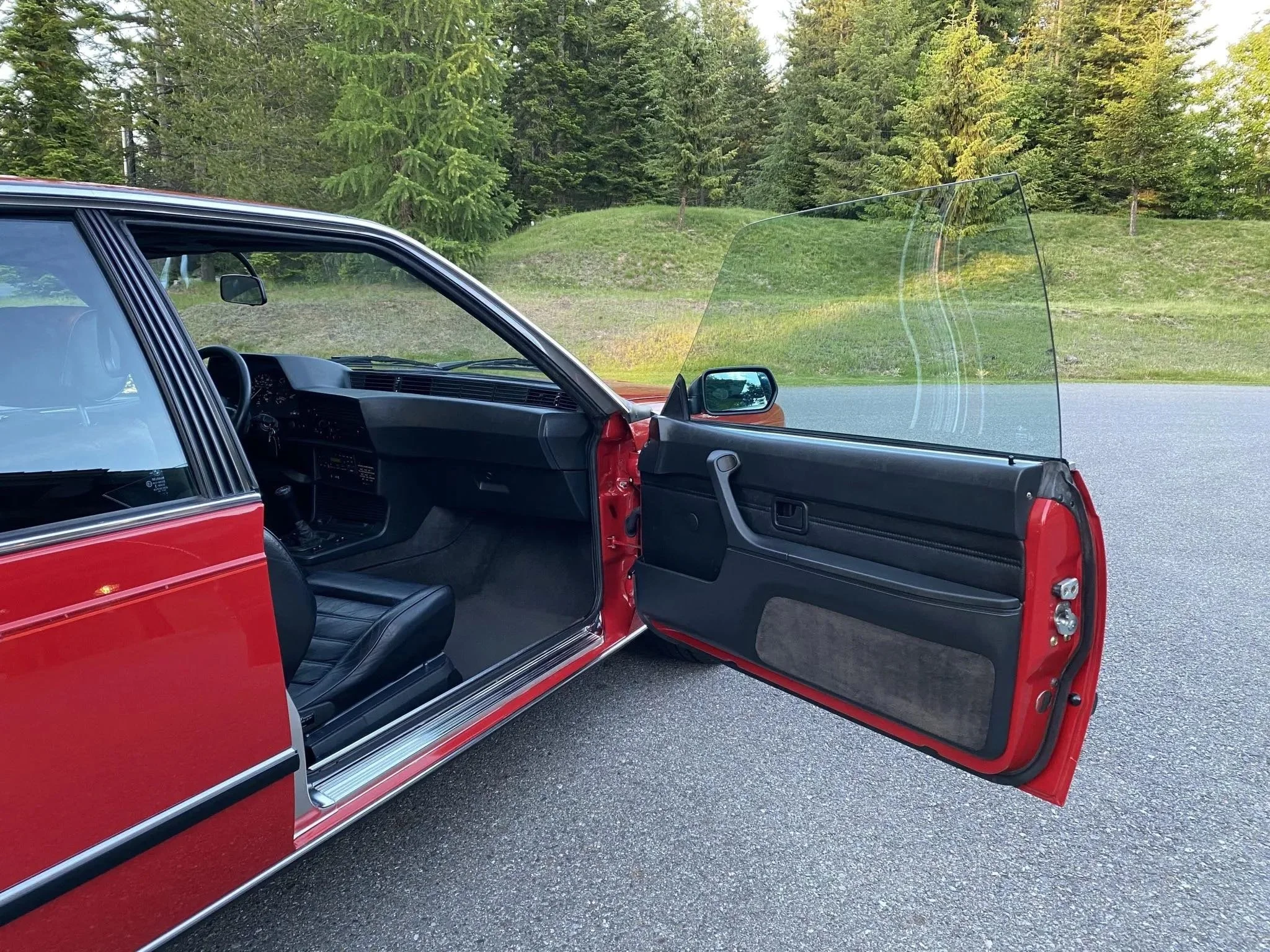 Open red vintage car door showing interior with black seats, dashboard, and a windshield with a clear view of trees and grass outside.