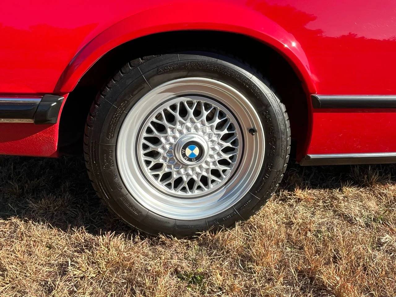 Close-up of a red classic BMW car wheel with a white mesh alloy rim, branded with the BMW logo in the center, parked on dry grass.