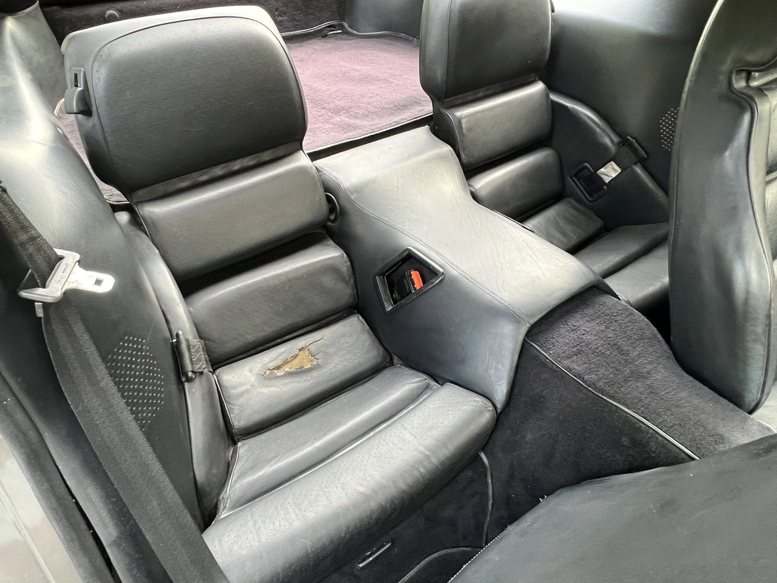 Rear seats of a car with black leather upholstery, including one seat with a tear in the material.