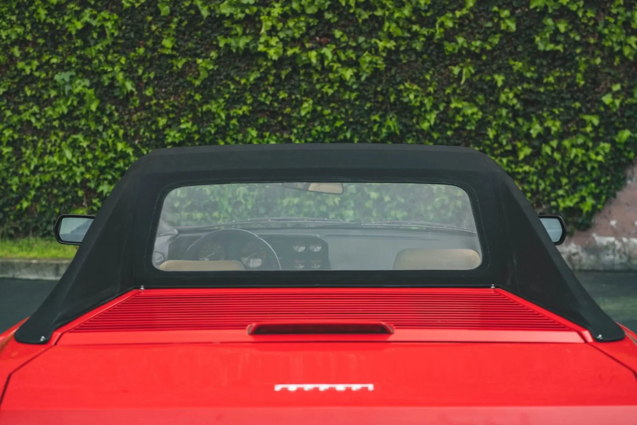 Red sports car with a black soft top roof, parked in front of a green hedge.