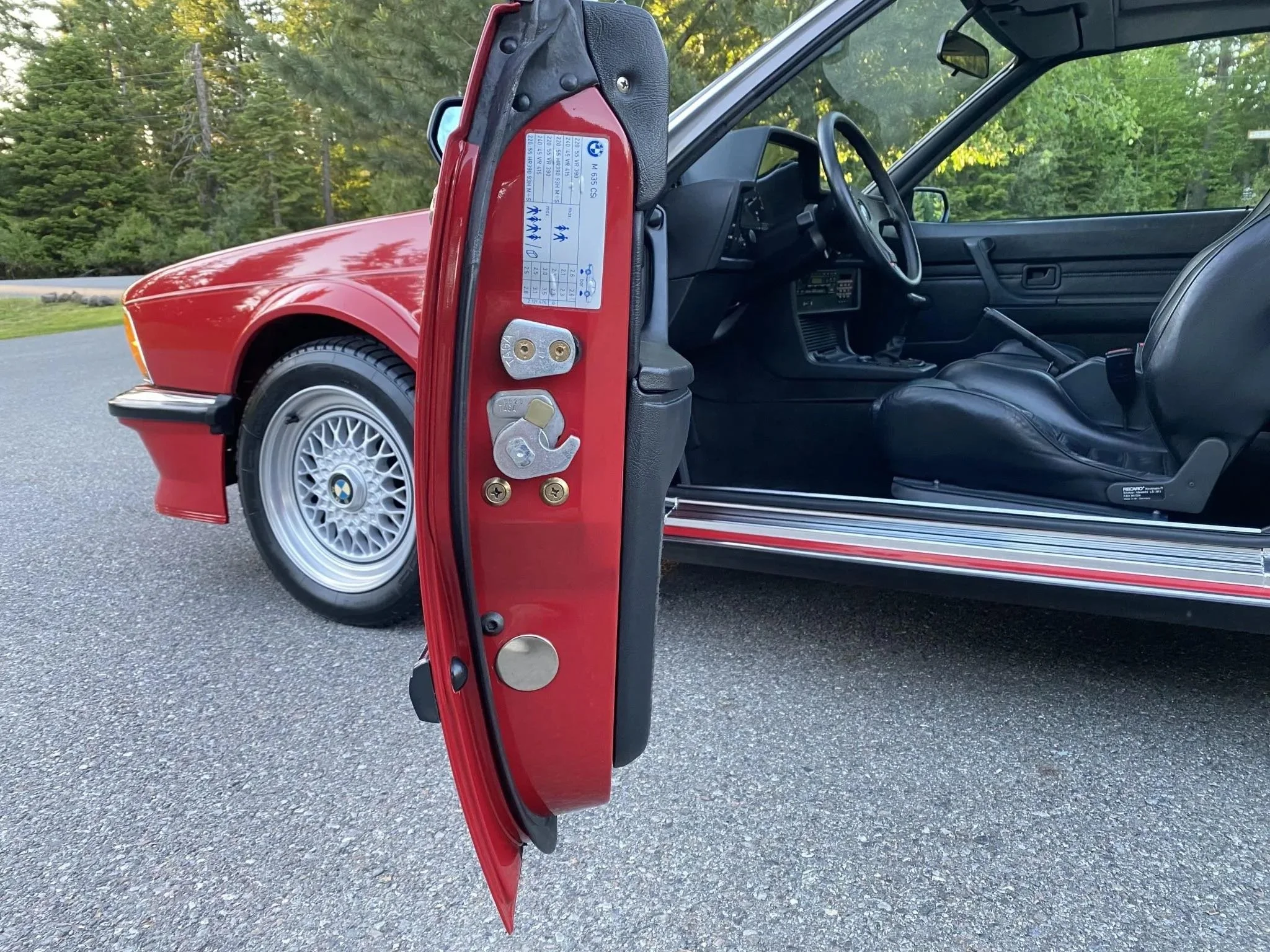 Close-up of the open driver side door of a red vintage BMW car with black interior and alloy wheels, parked on a paved road with greenery trees in the background.