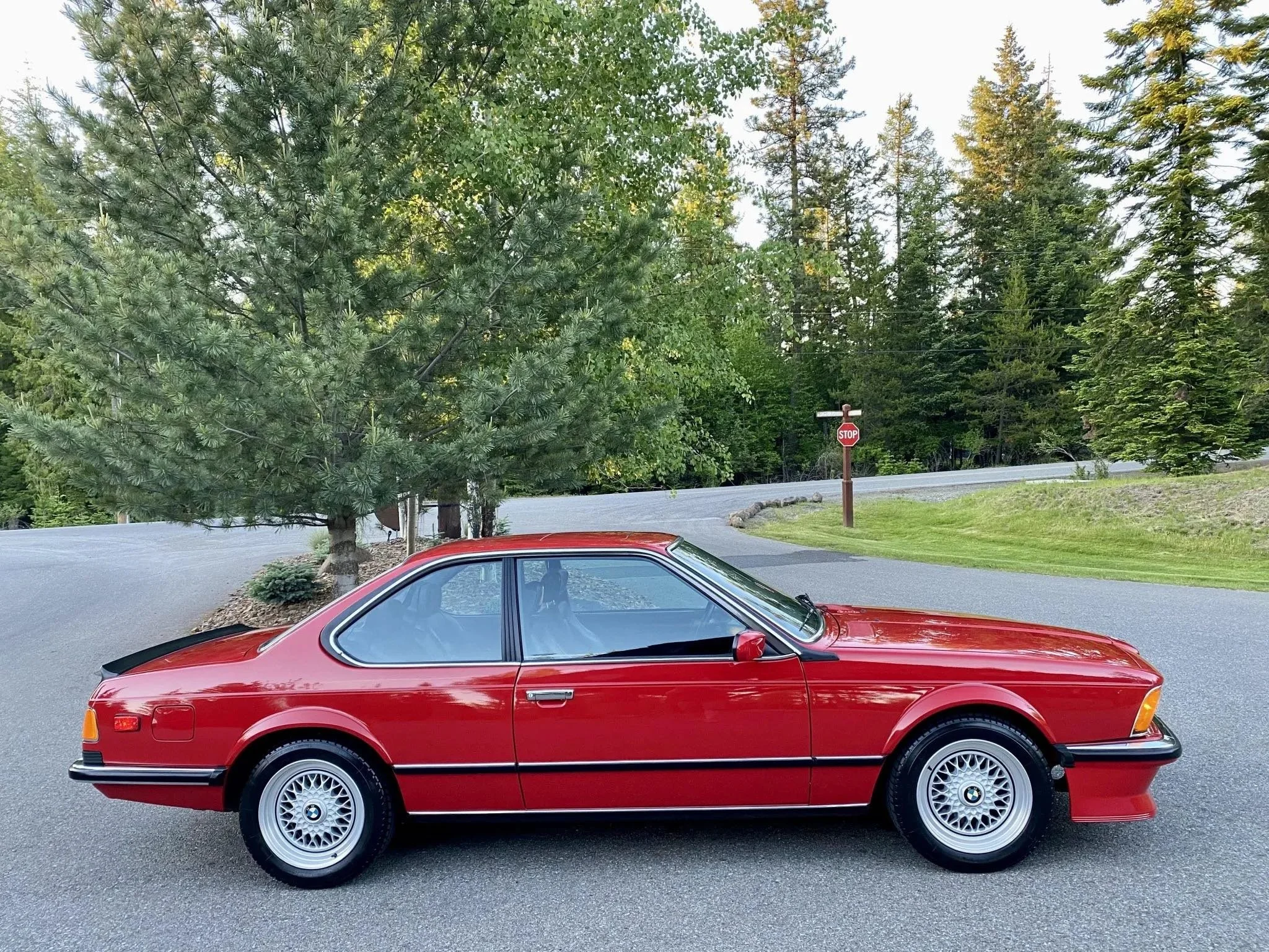 A red vintage BMW coupe parked on a paved road with trees and a stop sign in the background.