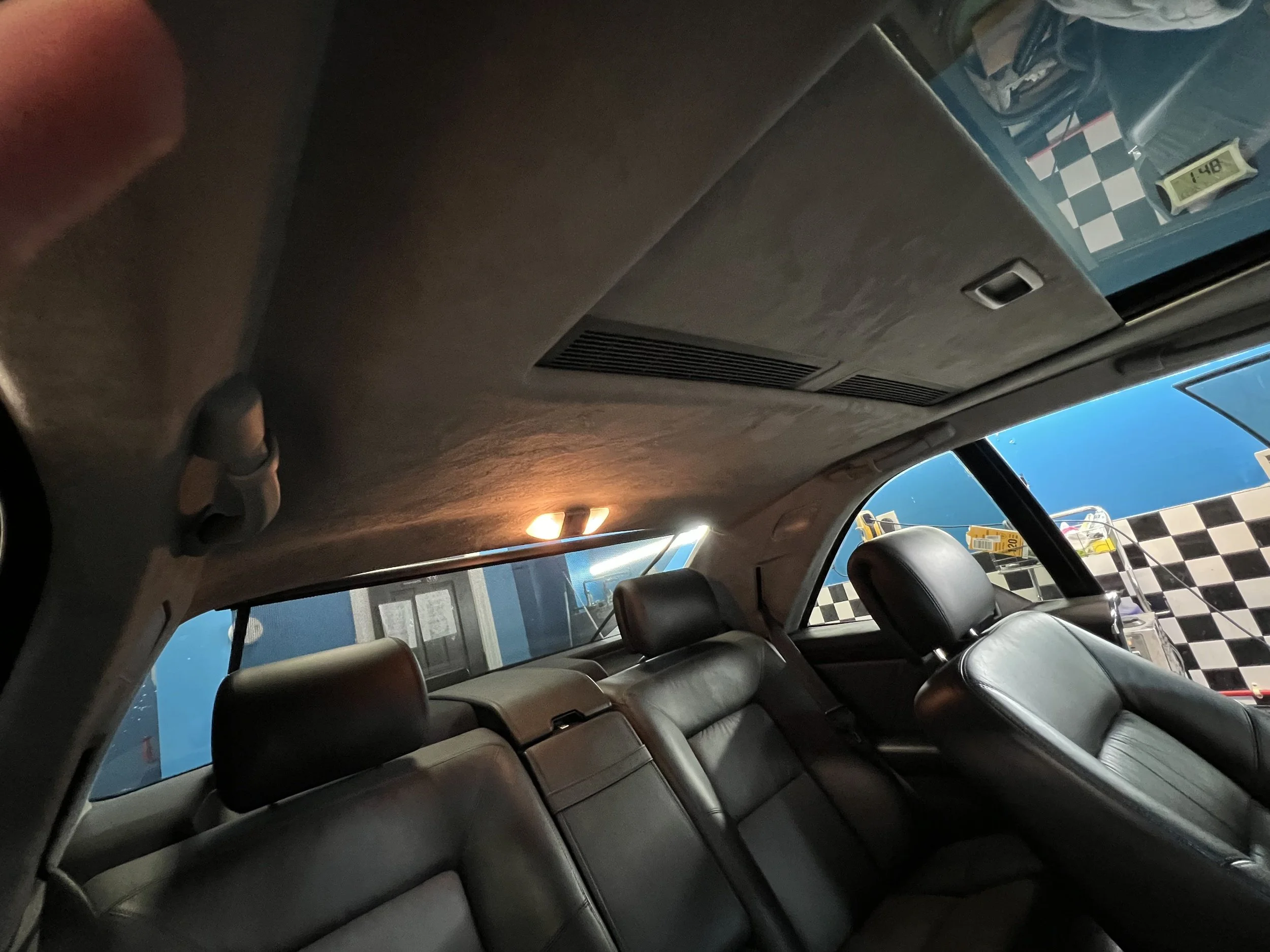 Interior view of a car showing black leather seats, a ceiling with a light, and a view through the rear window of a blue wall and checkered floor pattern in a garage or repair shop.