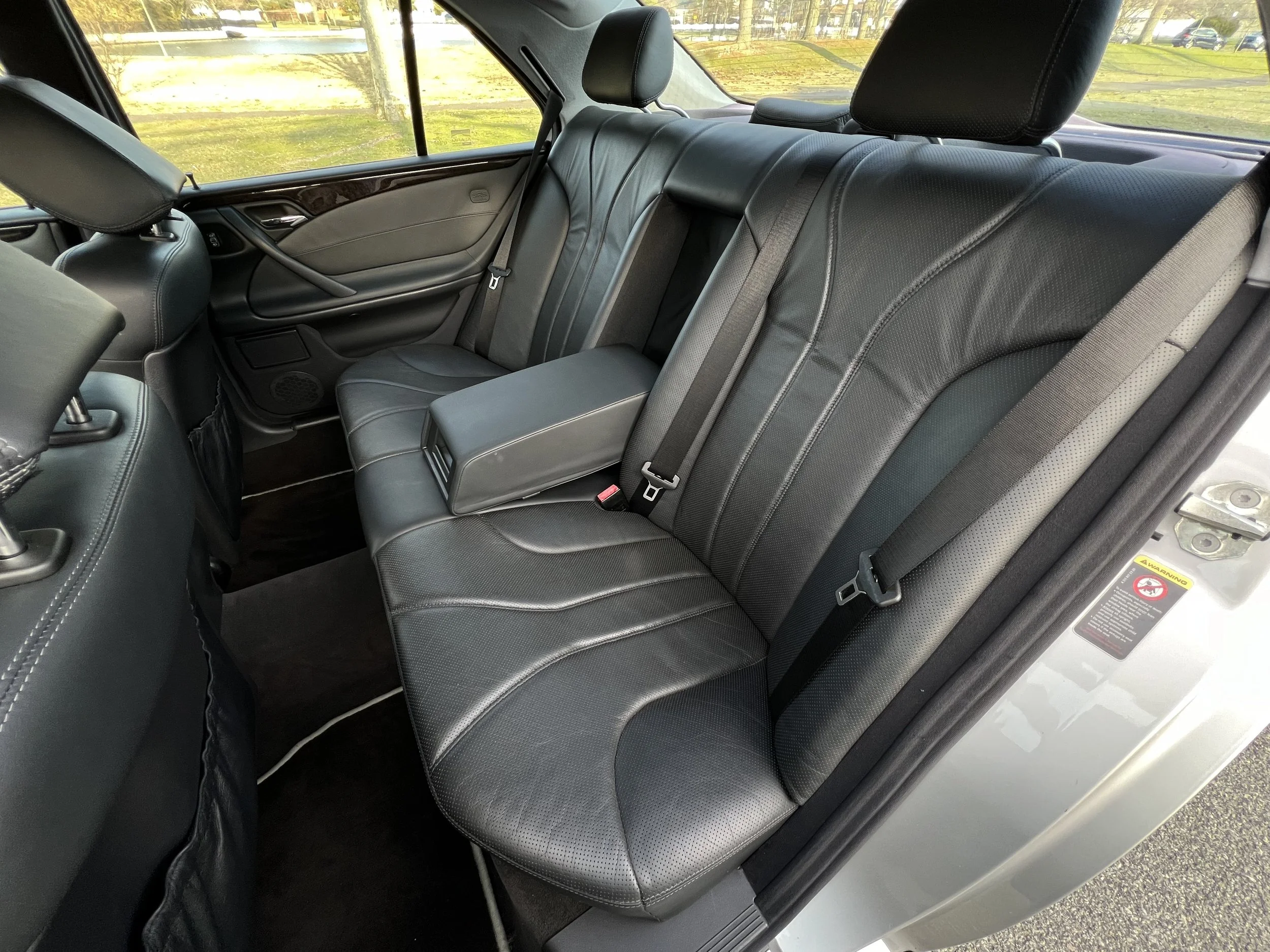 Back seat of a car with black leather seats, headrests, and seat belts, with a view of a grassy area and parked cars outside through the windows.