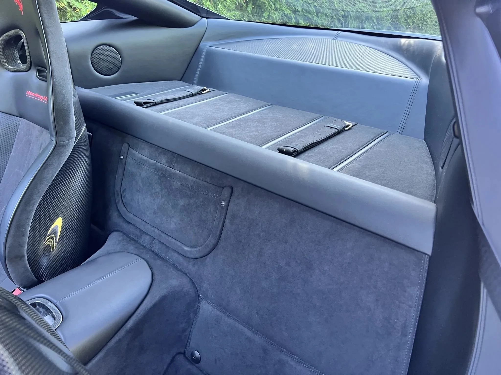 Interior of a car's rear section showing a flat cargo area with straps, a speaker, and upholstery panel.