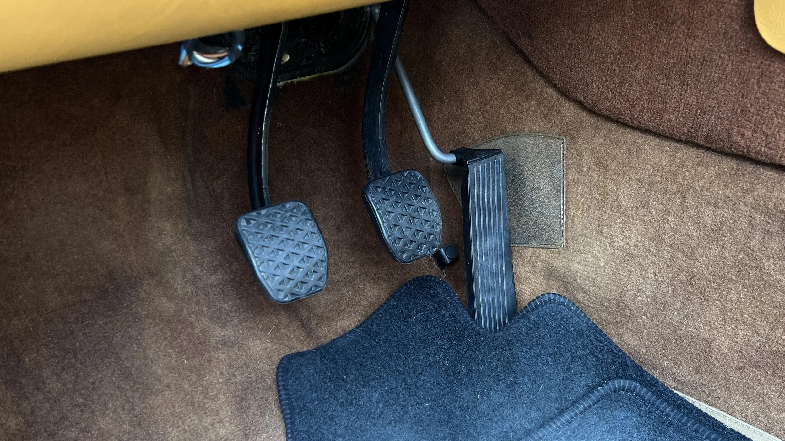 View of the brake and accelerator pedals in a 1977 BMW 630 csi, with a black floor mat and brown carpeting.
