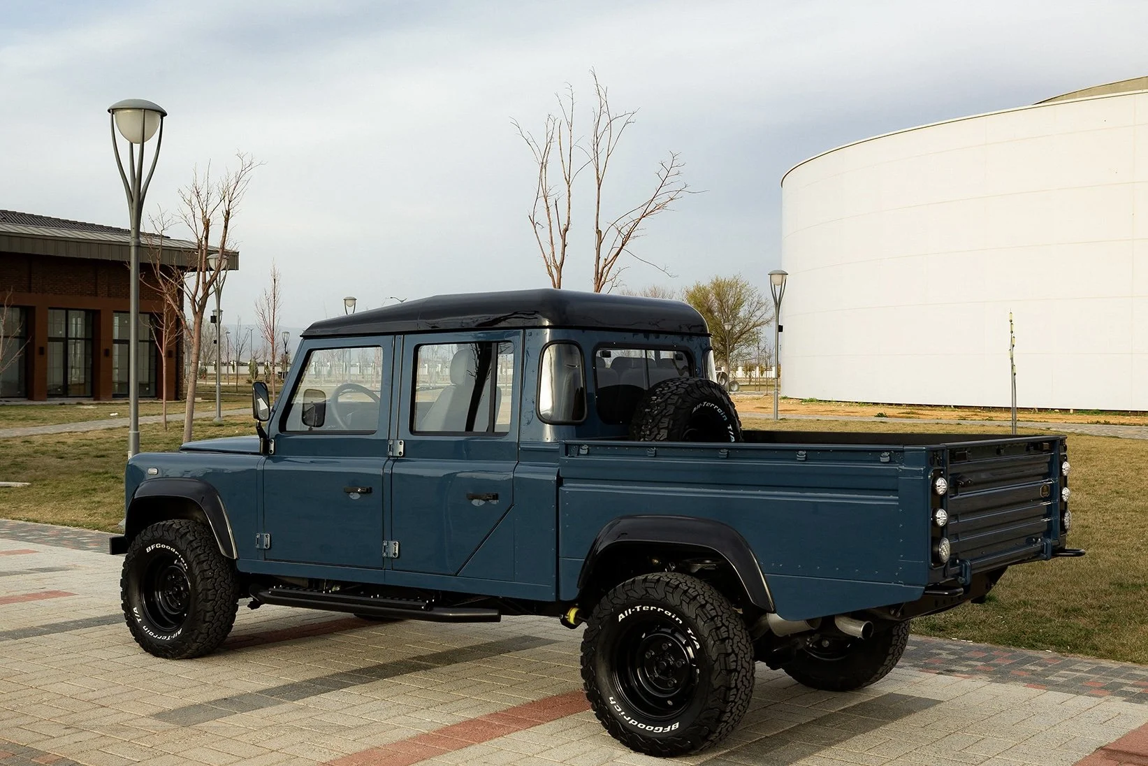 A vintage blue off-road vehicle, resembling a Land Rover Defender, parked on a paved area with a grassy background and modern buildings.