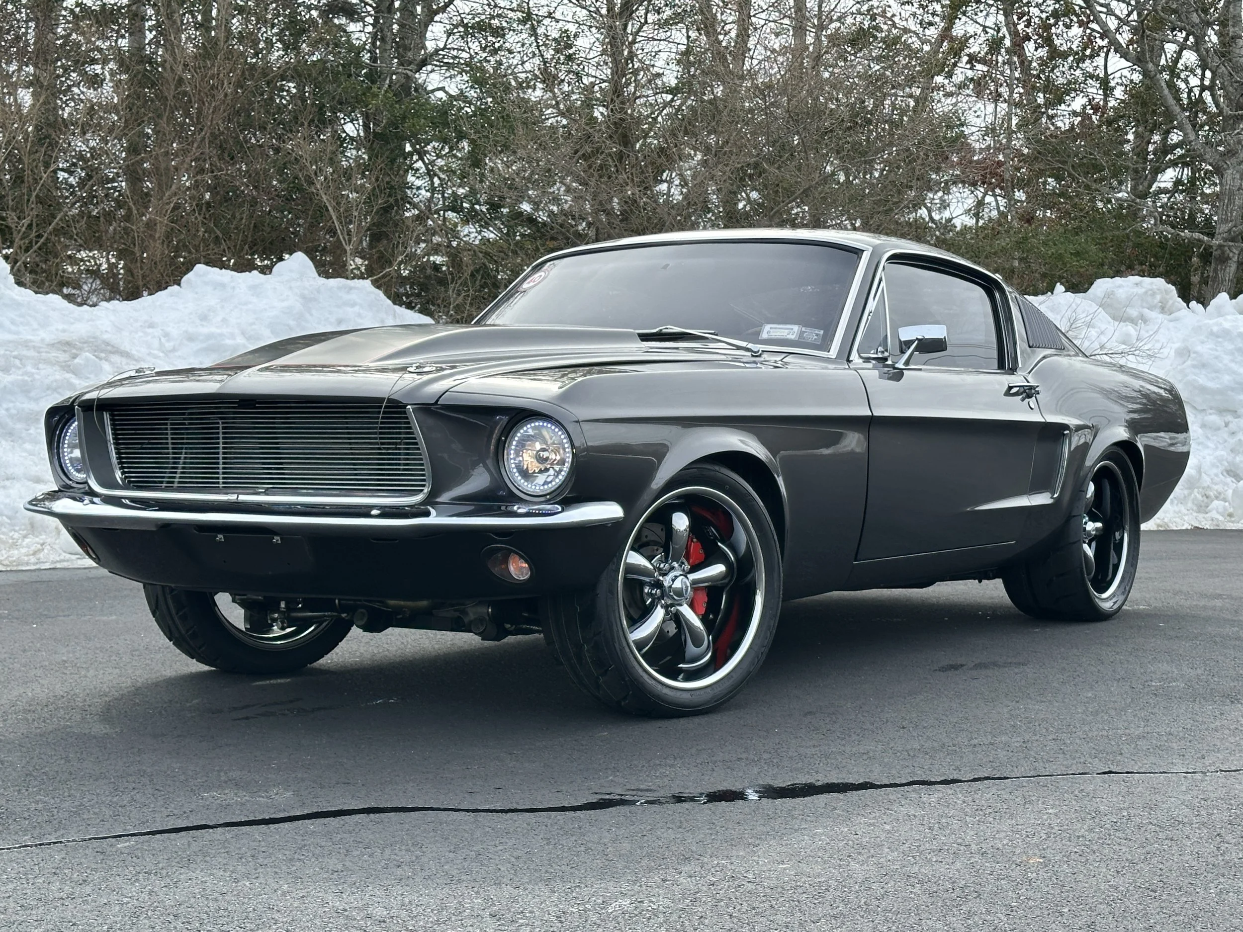 A black vintage Ford Mustang car parked on the pavement with snow piles in the background.
