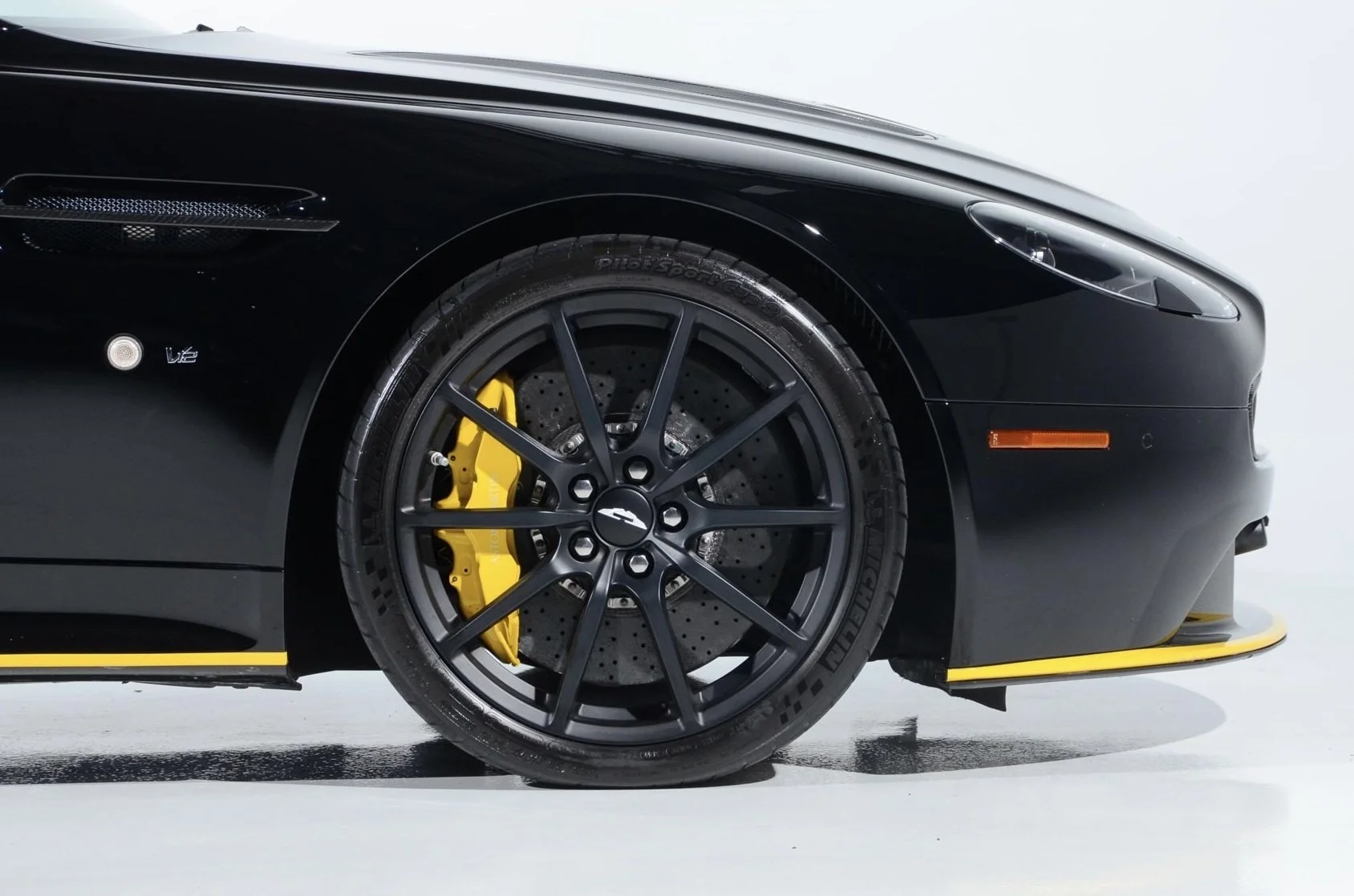 Close-up of a black luxury sports car front wheel with yellow brake caliper and perforated brake disc, part of the car's sleek body with vents and logo, on a white background.