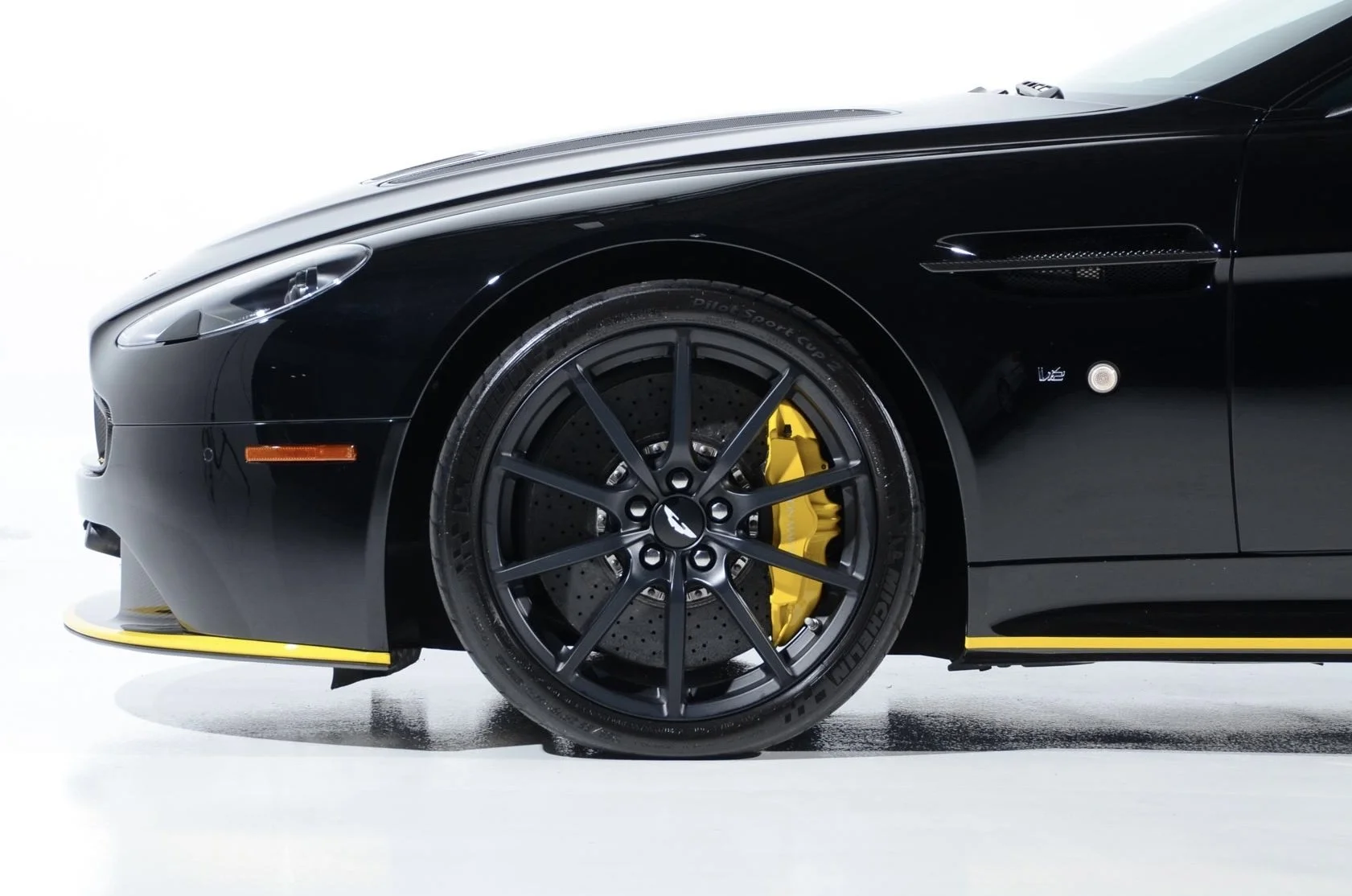 Close-up of the front wheel of a black luxury sports car with yellow brake caliper and matte black rim, featuring a Tesla logo at the center.