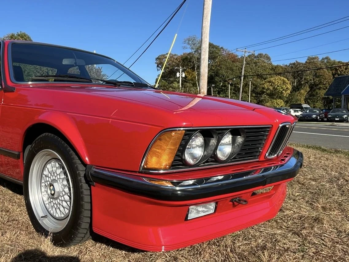 Red vintage BMW car parked on a grassy area near a road, with a clear blue sky and trees in the background.