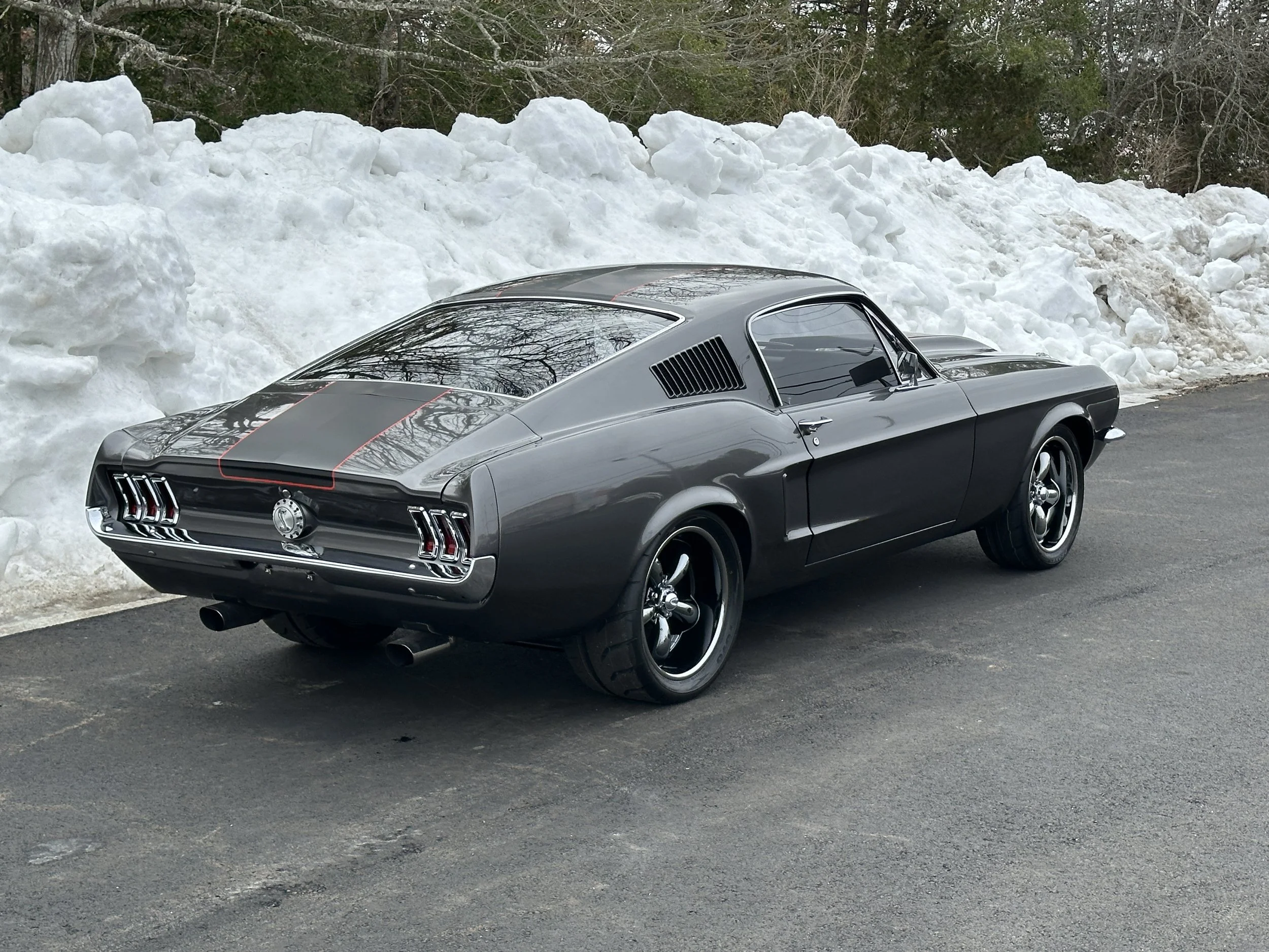 A vintage black and gray muscle car parked on an asphalt road next to a snowbank, with bare trees in the background.