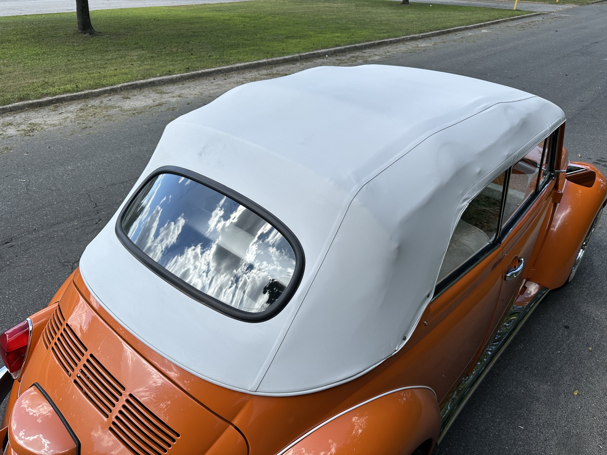 An orange vintage convertible car with a white soft top parked on a paved street, with grass and trees in the background.