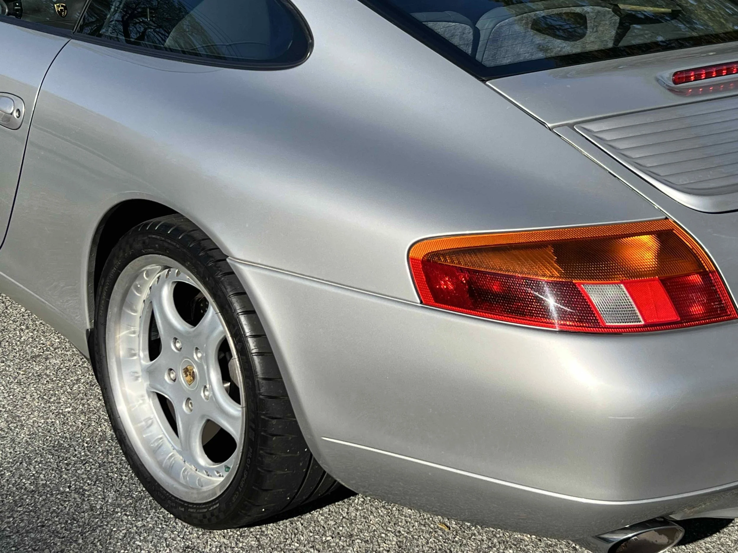 Close-up of a silver Porsche sports car showing part of the rear wheel, tail light, and rear quarter panel on a gravel surface.