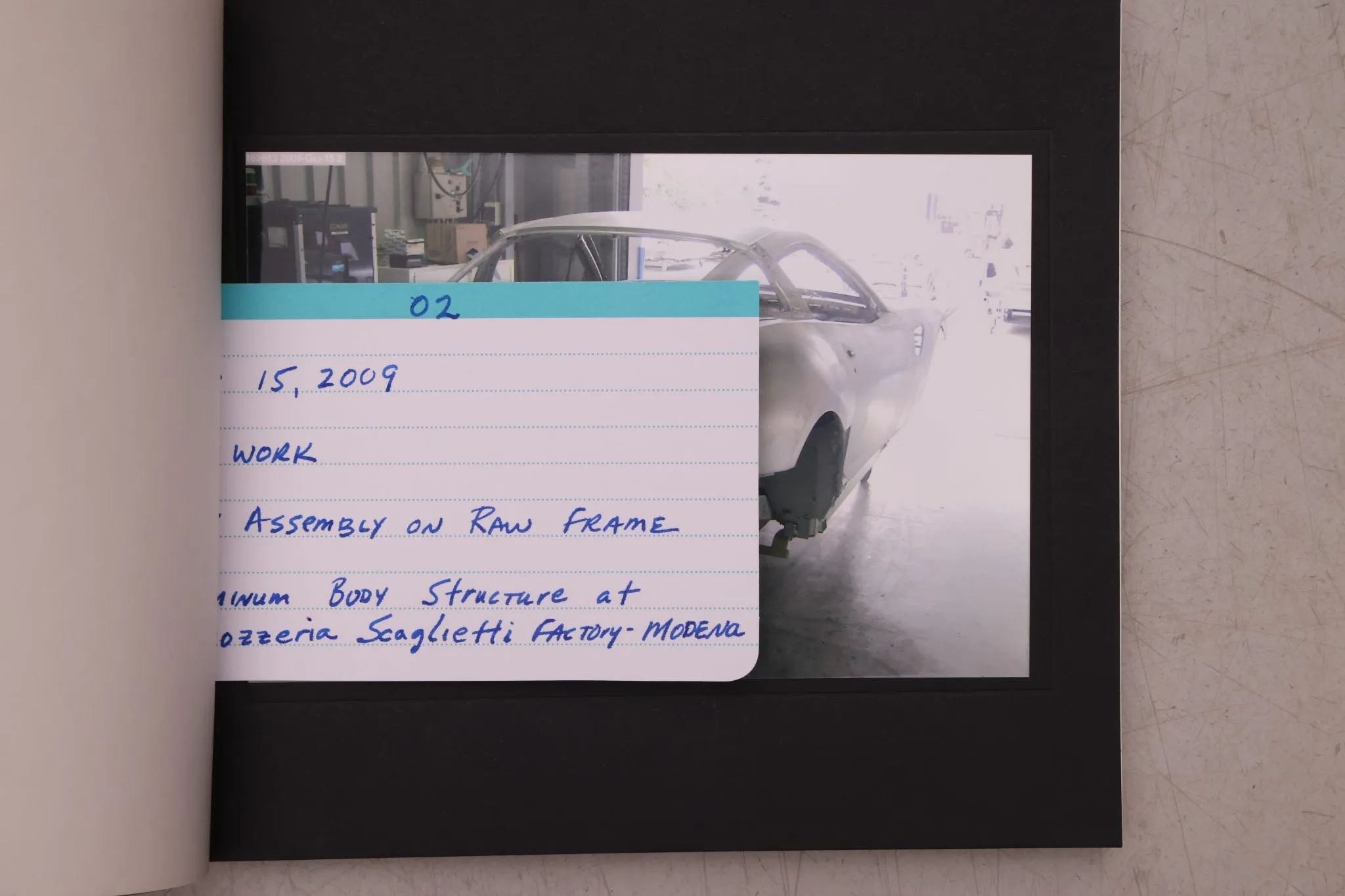 Photograph of a vintage car in a workshop with a handwritten note in front of it. The note details the date, work, and location of the assembly process on a raw frame at a car factory.