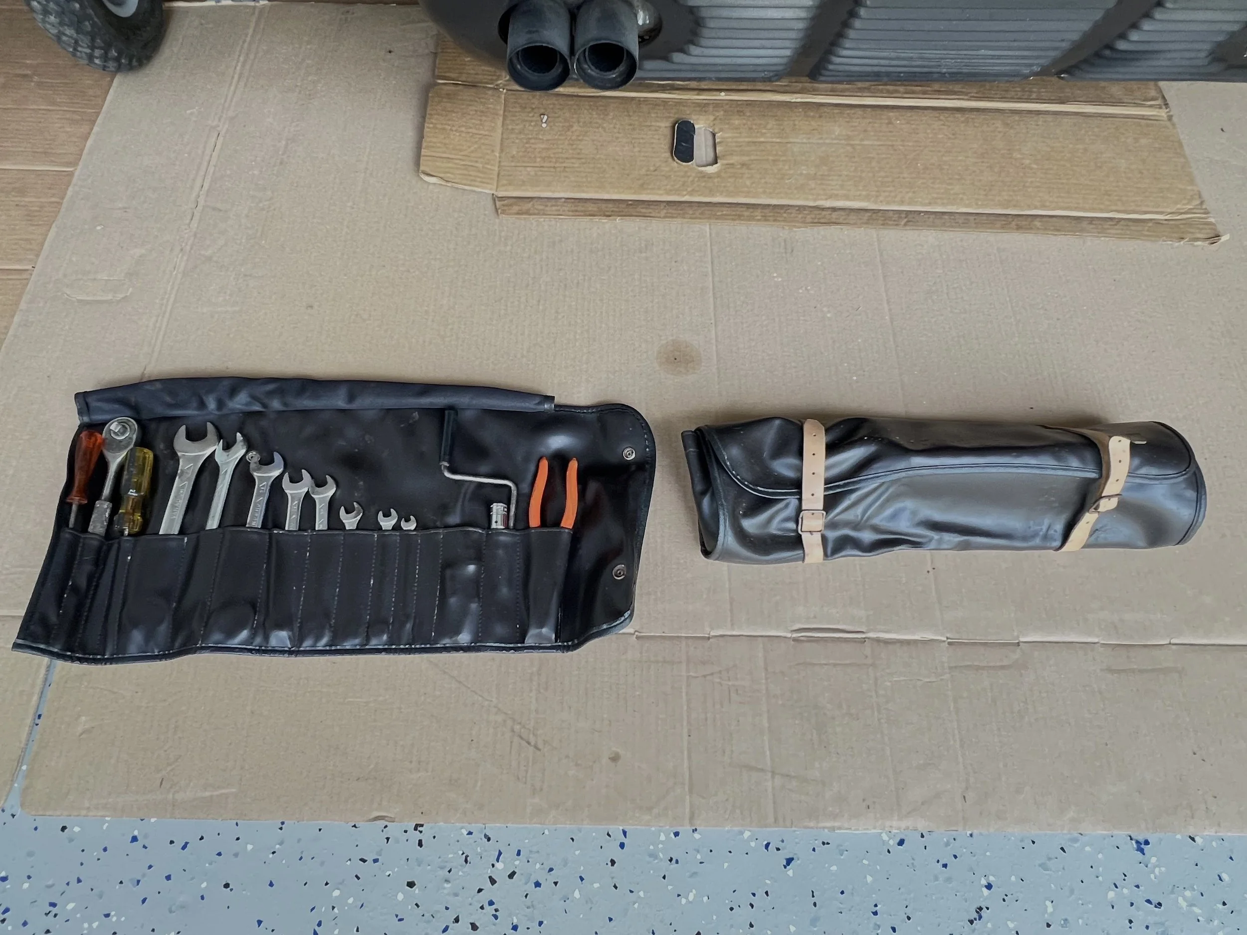 Tools laid out on a cardboard surface, including a black tool roll with various wrenches, screwdrivers, and pliers, and a black rolled-up carrying case.