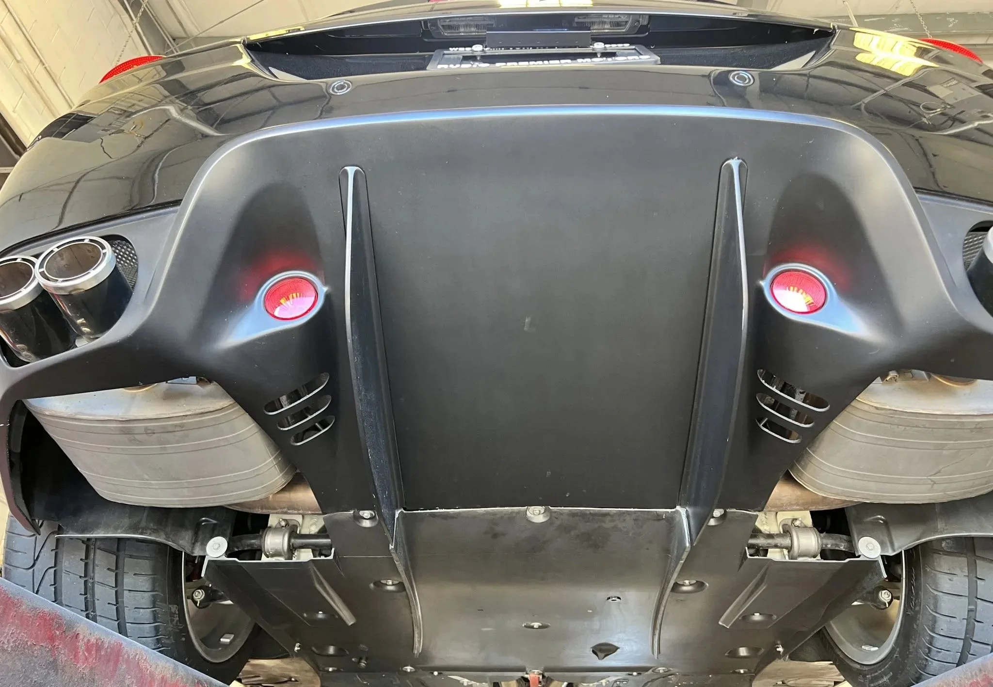 Underneath the rear of a black sports car, showing the exhaust pipes, suspension, and tires, with a custom protective panel covering the underside.