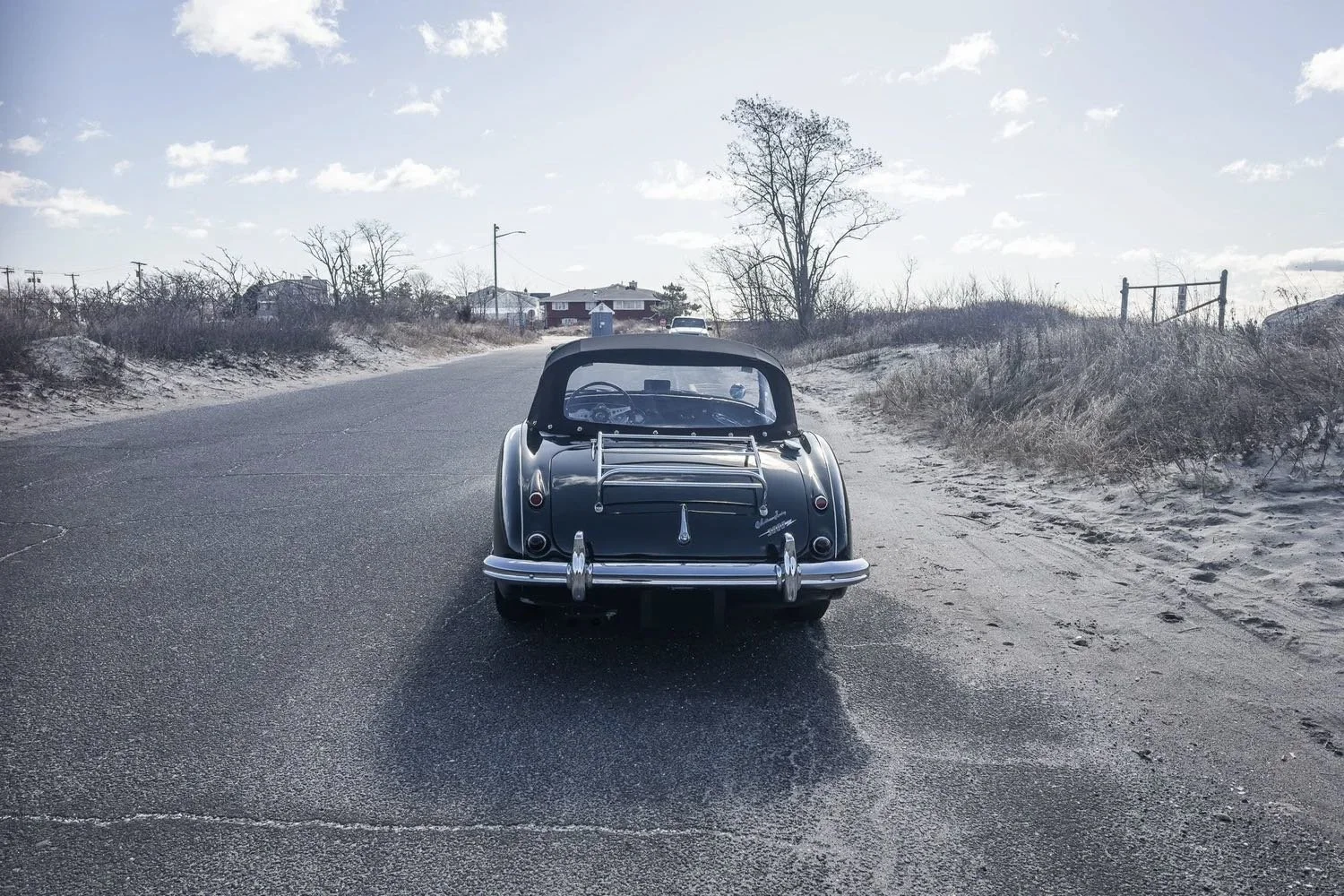 Rear view of a vintage car parked on the side of a rural road on a clear, partly cloudy day. The landscape includes dry sand and sparse trees.