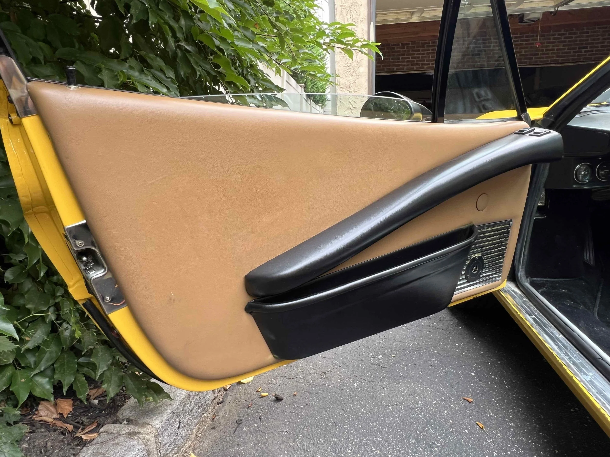 Close-up of a car door interior with beige upholstery, black armrest, black storage compartment, silver speaker grille, and a yellow exterior frame. Part of the car's black dashboard visible inside.