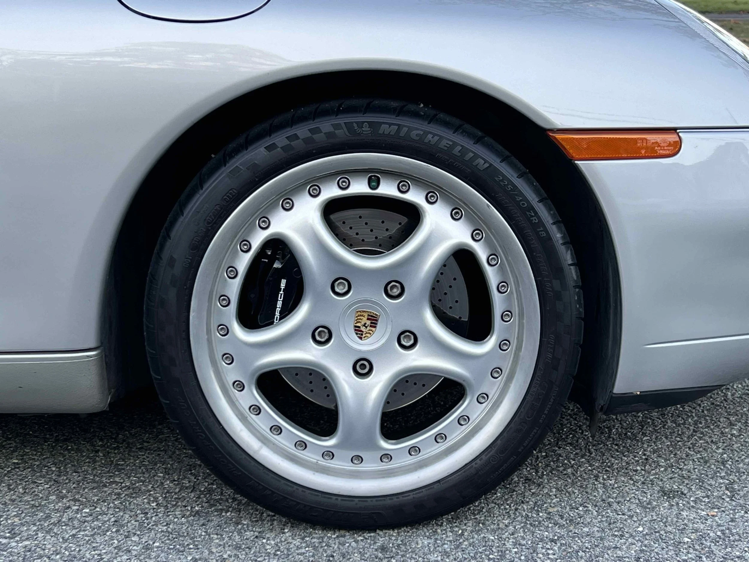 Close-up of a silver Porsche sports car front wheel with a Michelin tire, silver alloy rim, Porsche brake caliper, and side indicator light on a gray asphalt road.