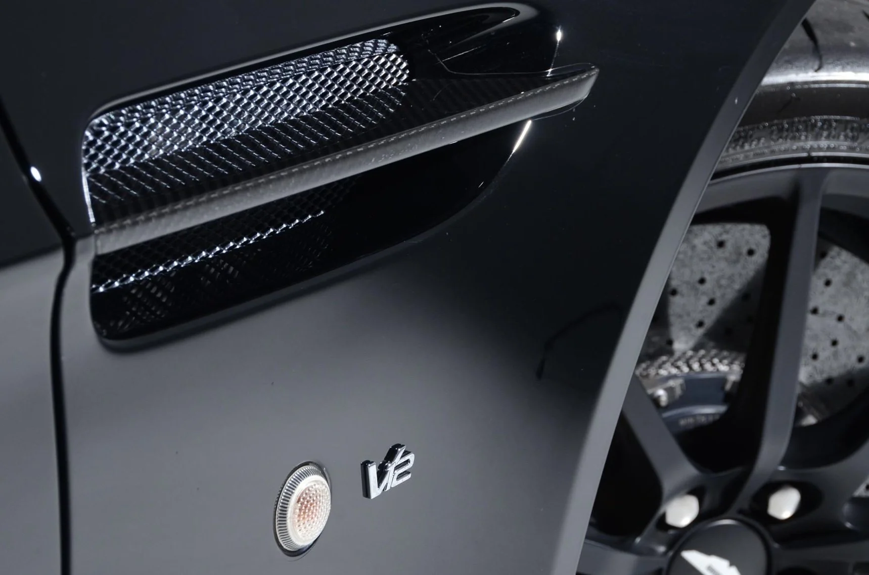 Close-up of a black car's front fender showing the side vent with a mesh grille, a circular turn signal light, and a 'V12' badge. Part of the wheel and tire is visible.