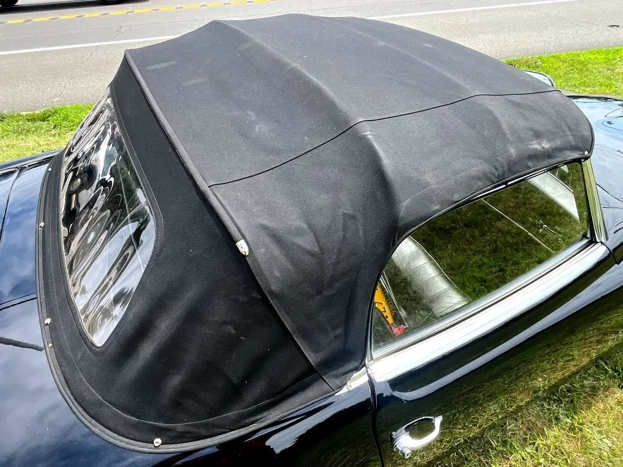 A classic car with a black soft top convertible roof, parked on grass next to a street.