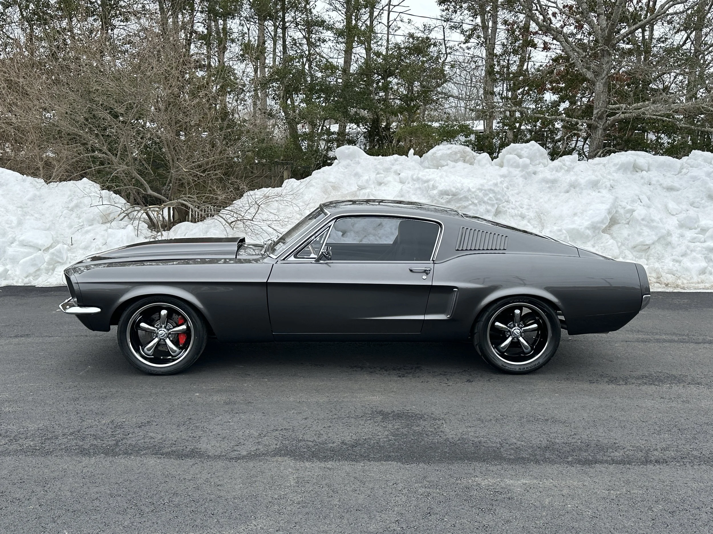 A black vintage car parked on a paved road with large snowbanks behind it and trees in the background.