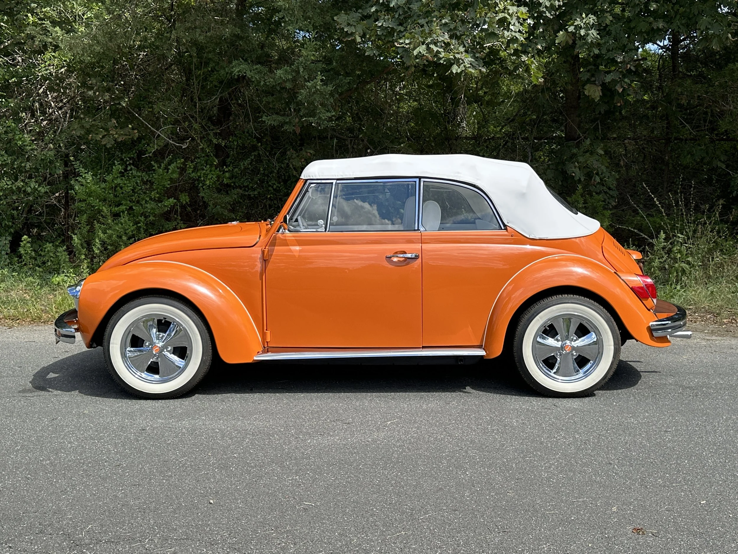 An orange vintage Volkswagen Beetle with a white convertible top parked on the road near trees.