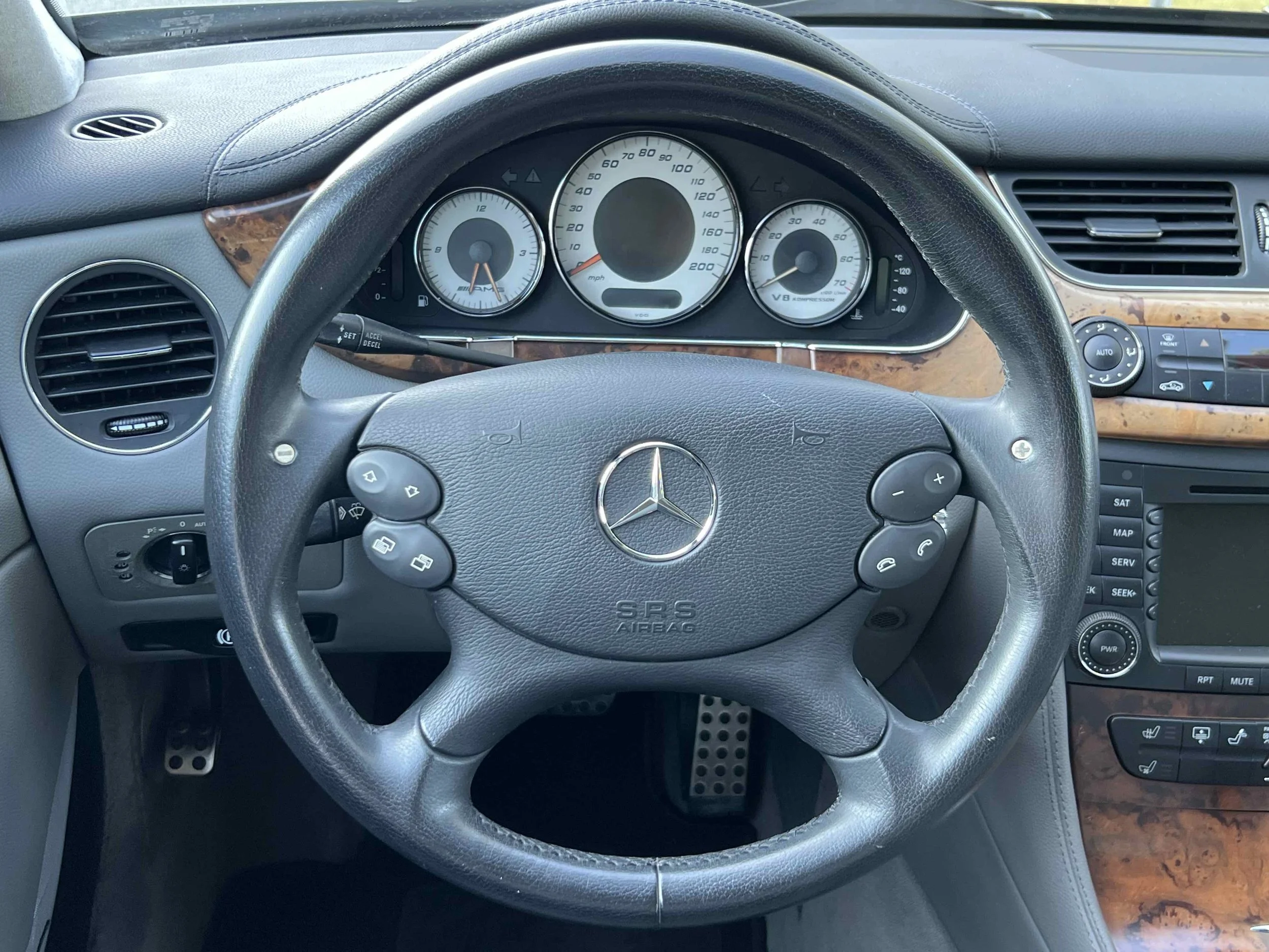 Interior view of a Mercedes-Benz vehicle dashboard and steering wheel, showing the instrument cluster, cruise control stalk, air vents, and center console with controls and wood trim.