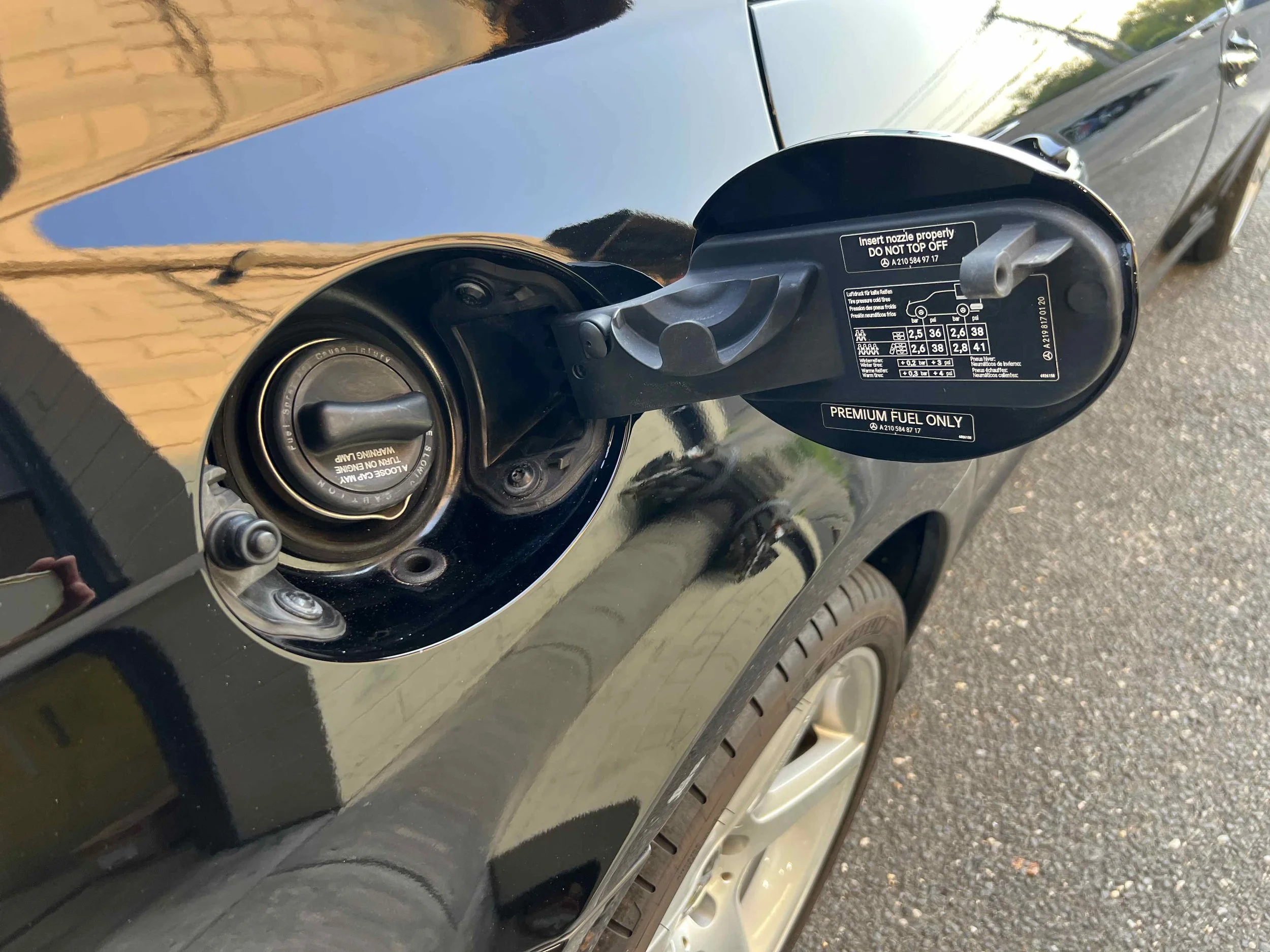 Car's fuel cap area with the fuel door open, showing a warning label and fuel type, with part of the tire and body of the car visible.