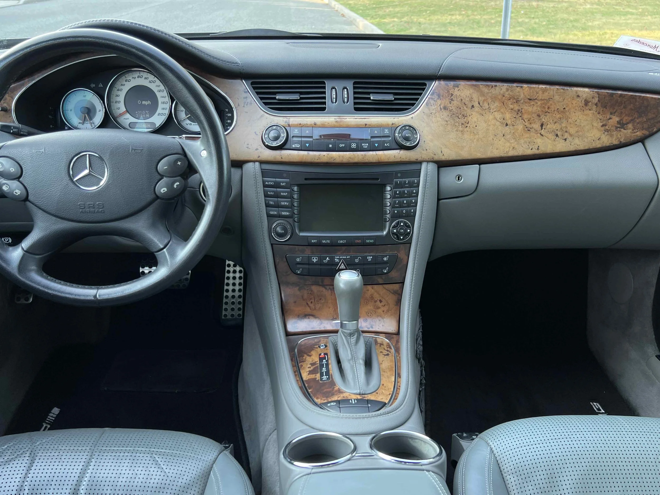Interior of a luxury car showing the dashboard, steering wheel with Mercedes-Benz logo, wood trim, center console with gear shift, and two cup holders.