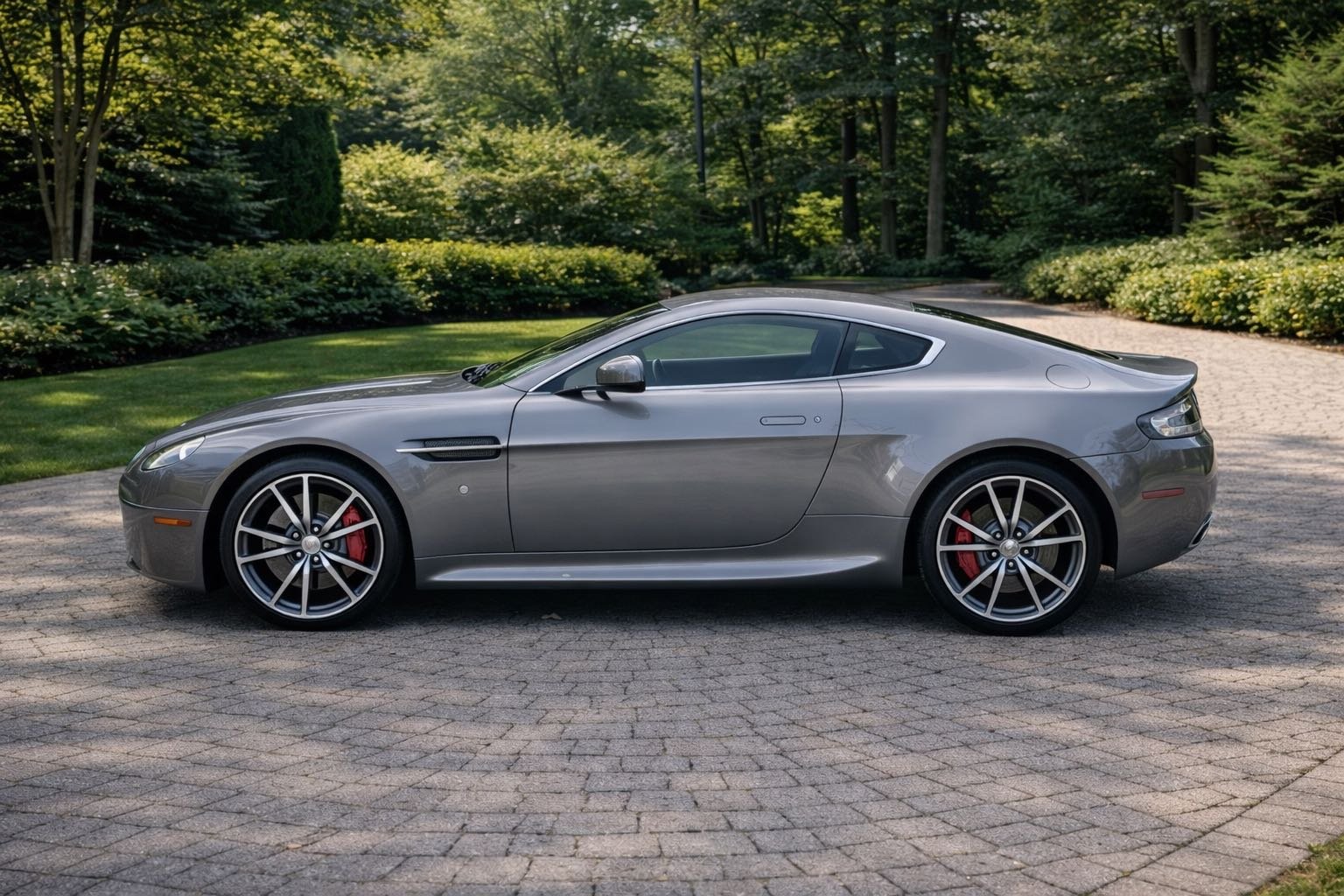 A silver sports car parked on a brick driveway with trees and green bushes in the background.