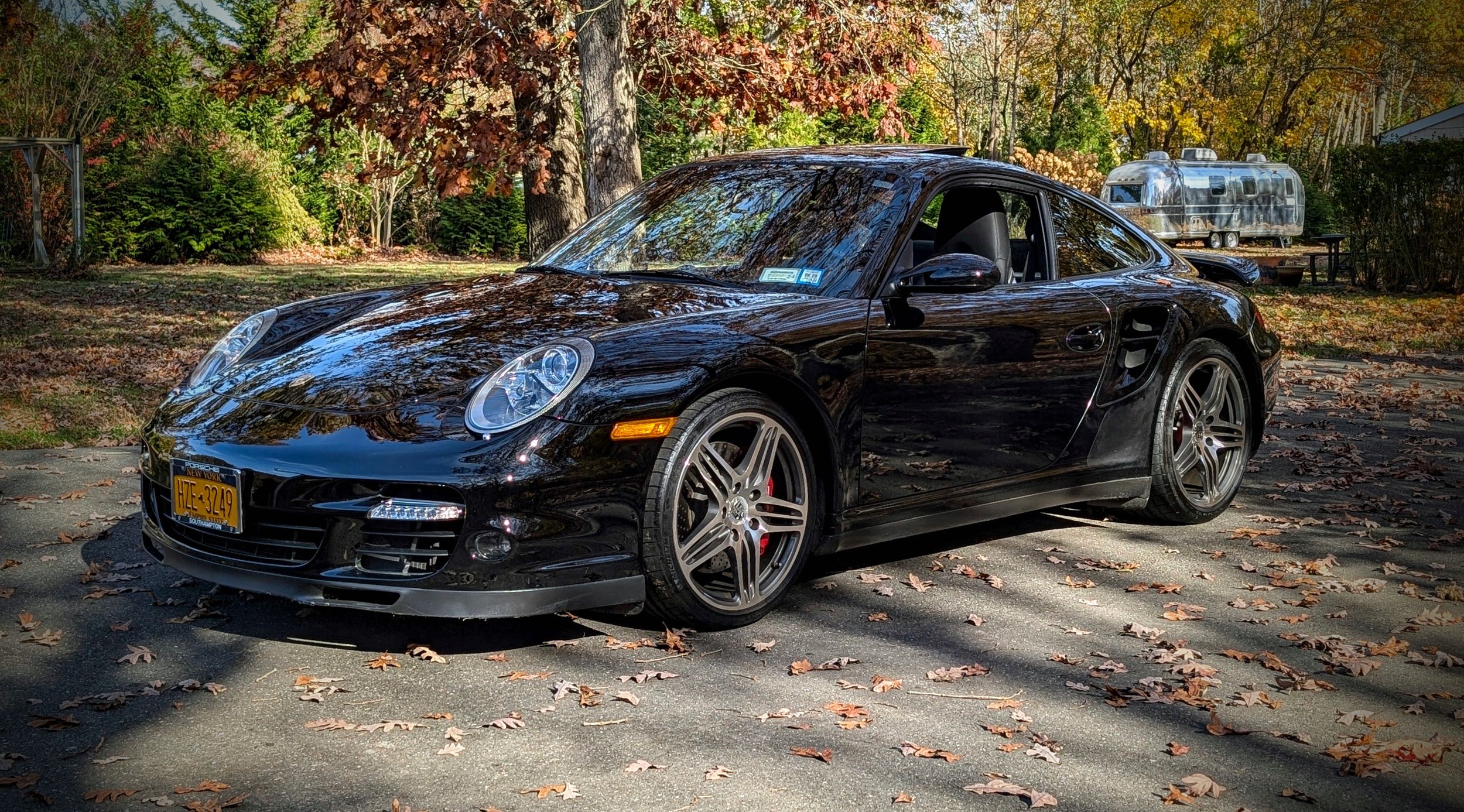 Black sports car parked on a street covered with fallen leaves in an autumn setting.