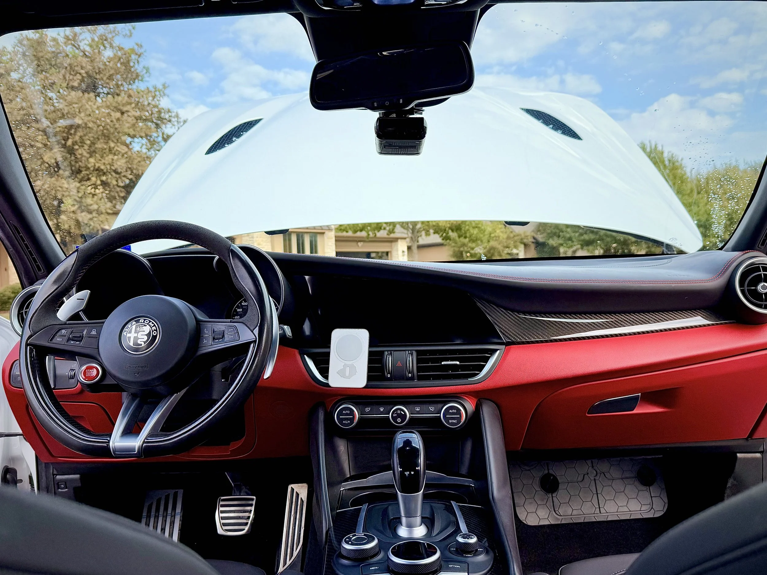 Inside view of a luxury sports car dashboard, showing steering wheel with Alfa Romeo logo, central console with gear shifter, climate controls, a mounted white phone holder, and a large windshield with an open hood of the car visible outside.
