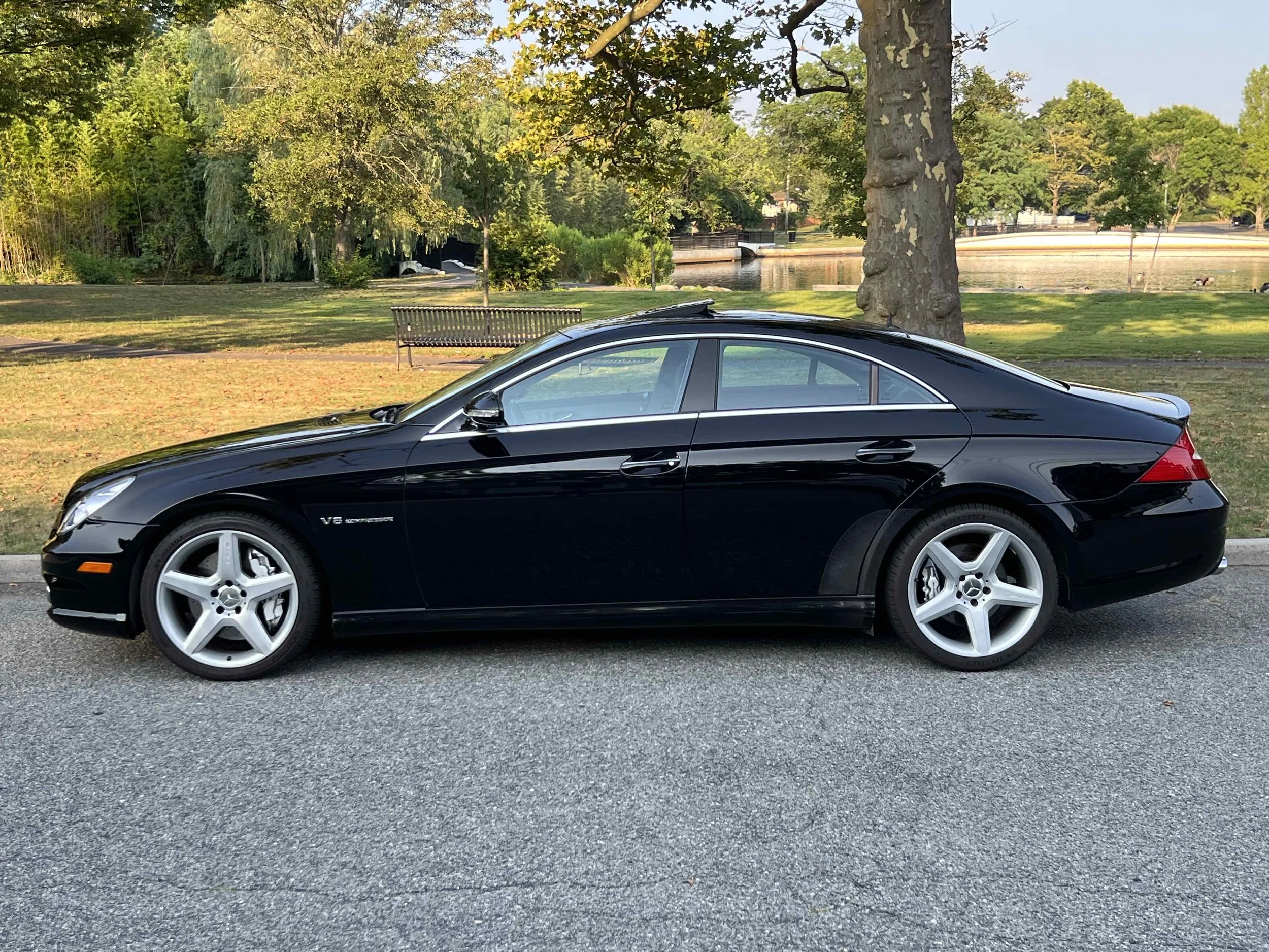 Black Mercedes-Benz CLS55 AMG  parked on a street near a park with grass, trees, a water body, a bench, and a bridge in the background.
