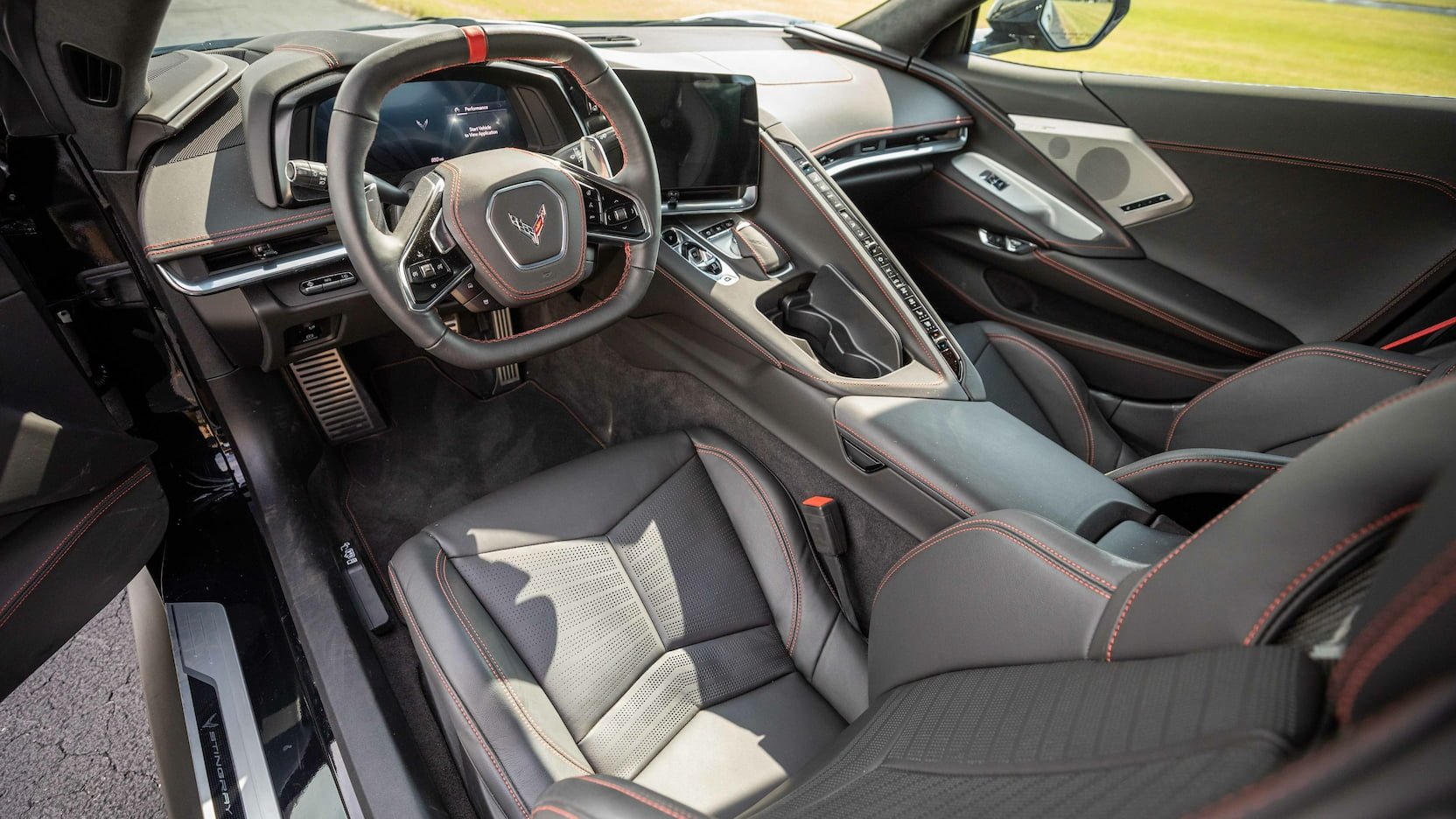 Interior view of a modern sports car showing the steering wheel, dashboard, center console, and front seats with black upholstery and red stitching.