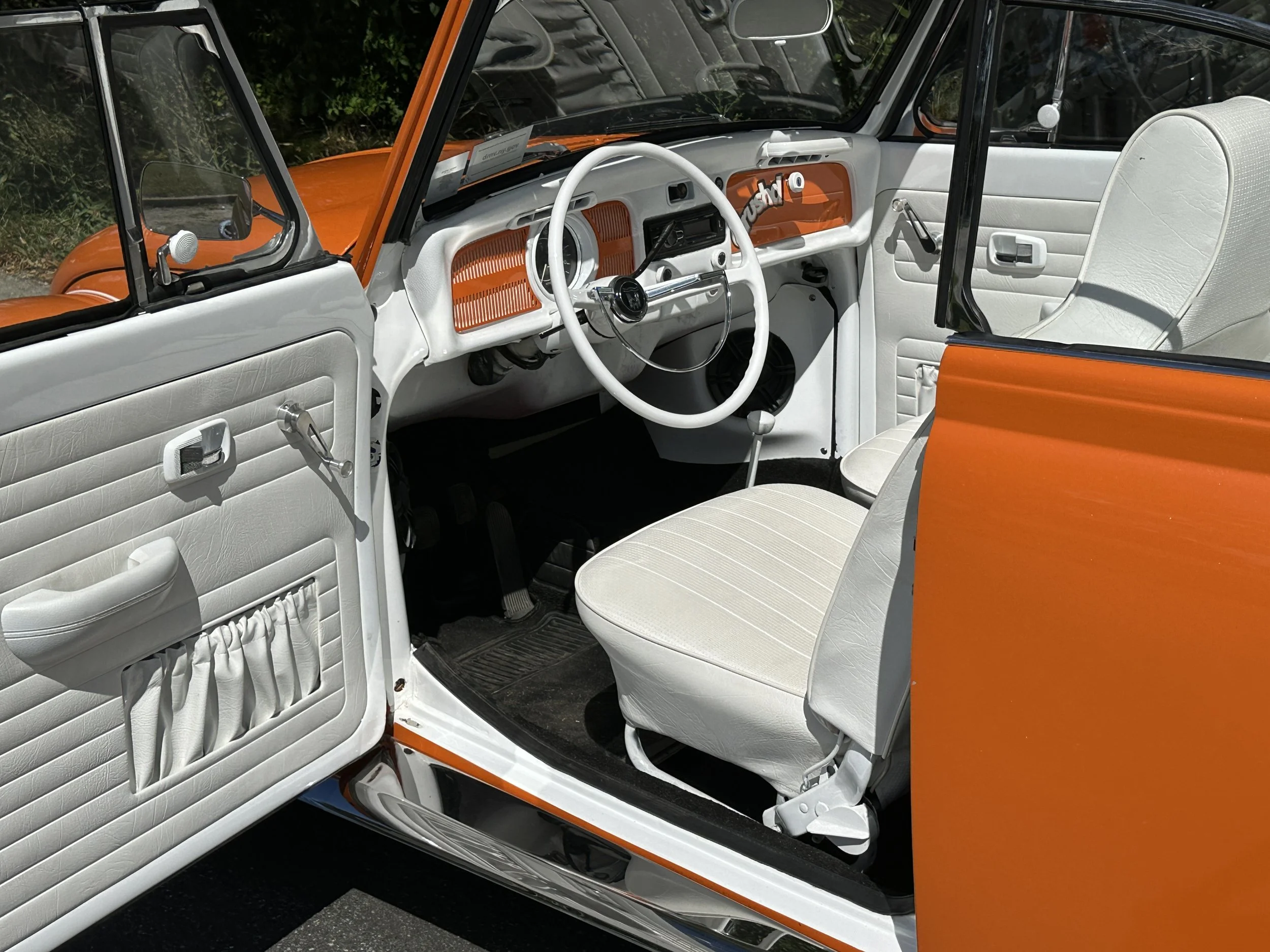 Interior of a vintage car with white seats, steering wheel, and dashboard featuring orange accents, with the driver's side door open showing a white door panel with storage pocket and window crank, and the exterior of the car in orange and white.